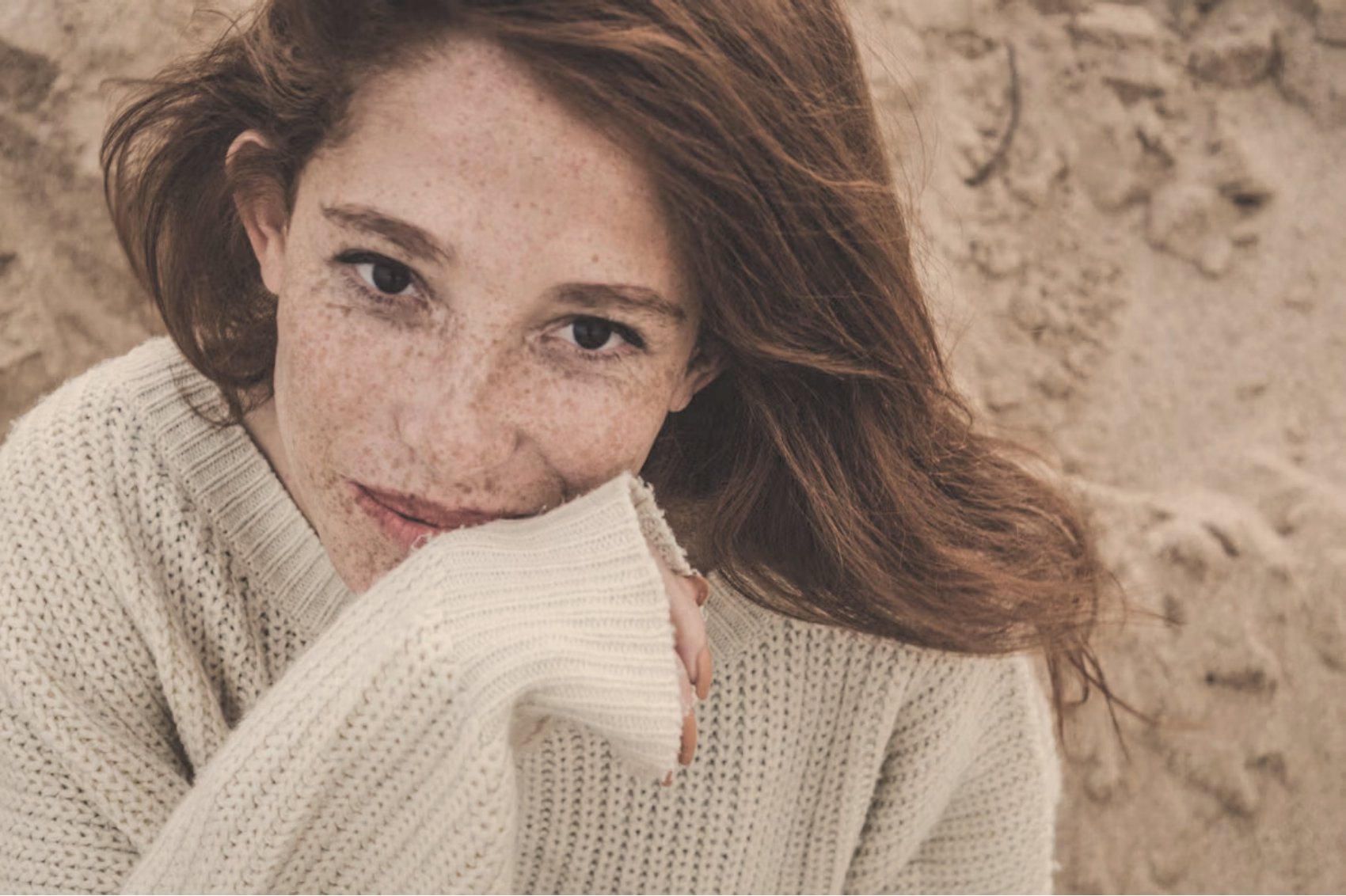 Close-up portrait of a freckled woman in a cream knit sweater against a textured beige wall