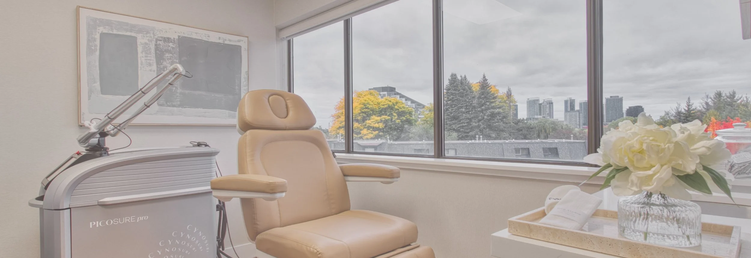 Modern aesthetic treatment room with a beige treatment chair, PicoSure Pro laser device, flowers, and city skyline views through large windows