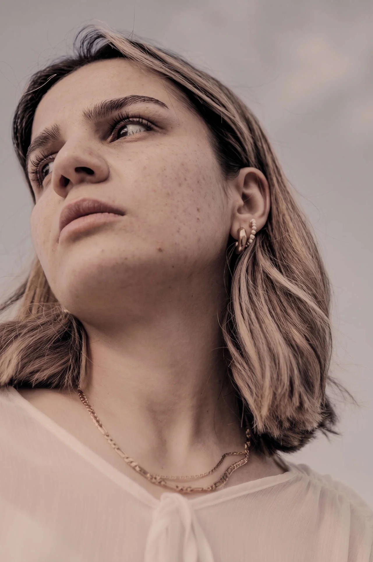 Close-up portrait of a woman with highlighted hair, freckles, gold earrings, and layered necklaces