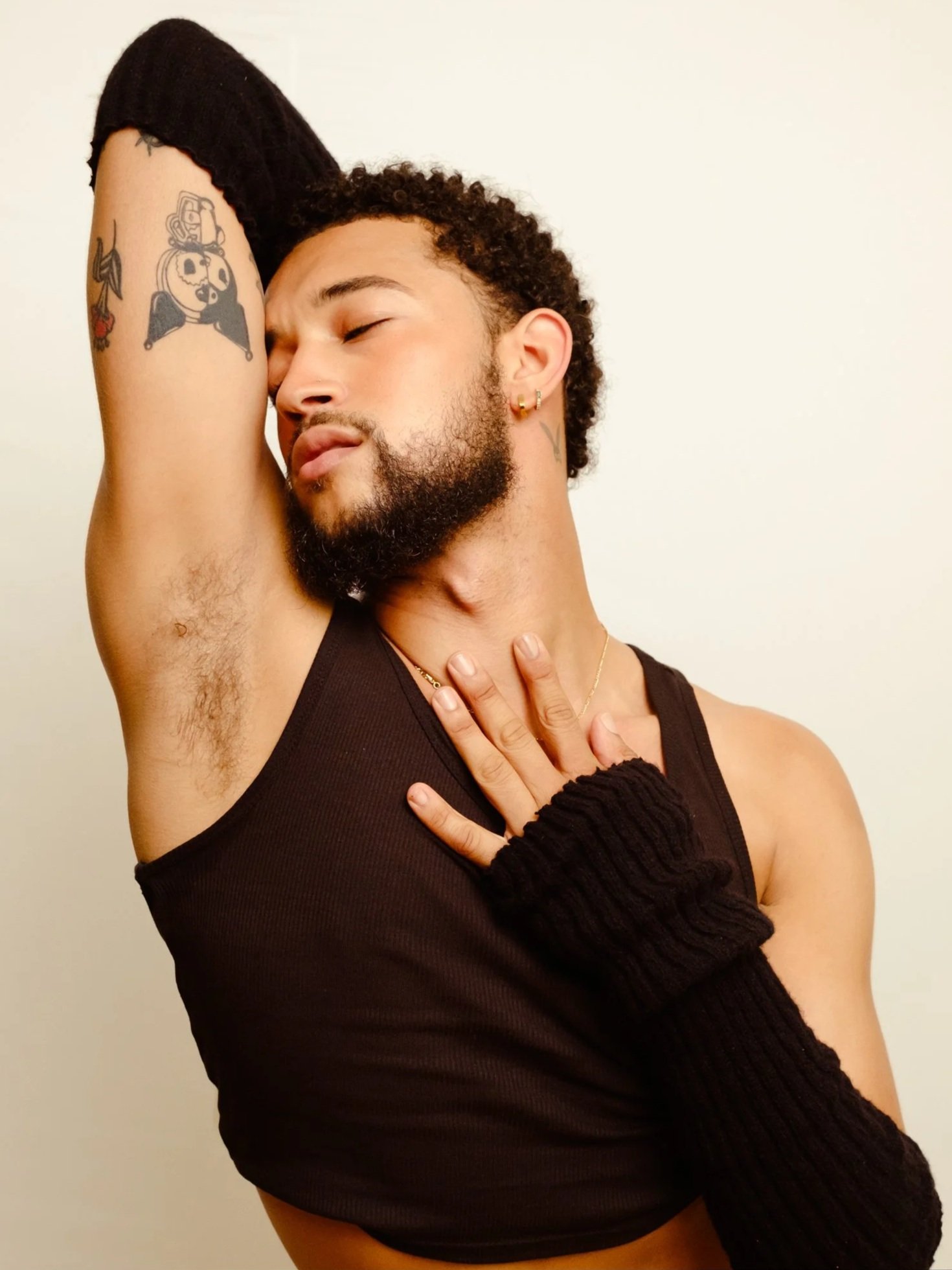 A person with curly hair and tattoos resting their forehead and arm against another person's armpit with closed eyes, wearing a black sleeveless top.