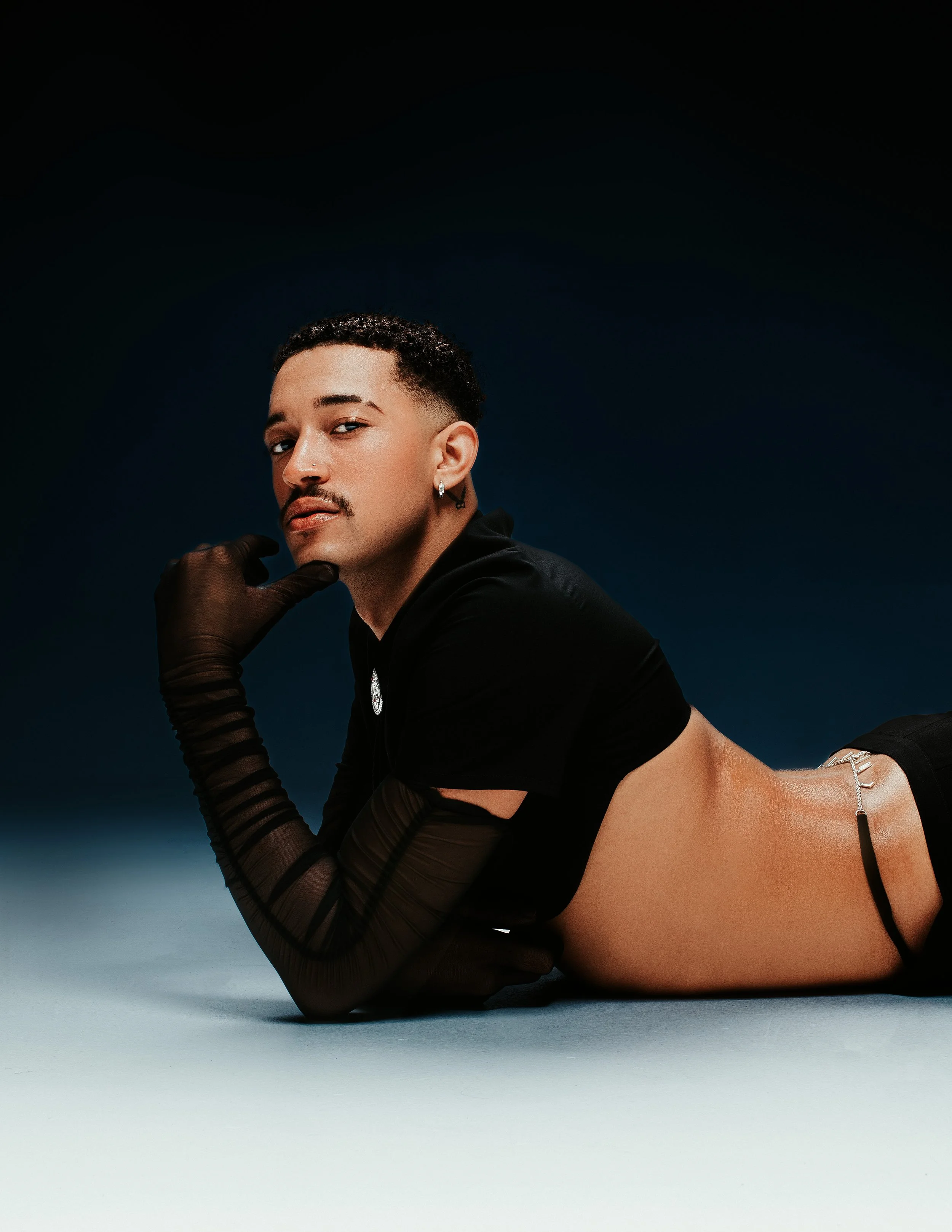 A person with short curly hair, a nose piercing, and earrings, lying on the ground, wearing black sheer gloves and black clothing, with a dark background.