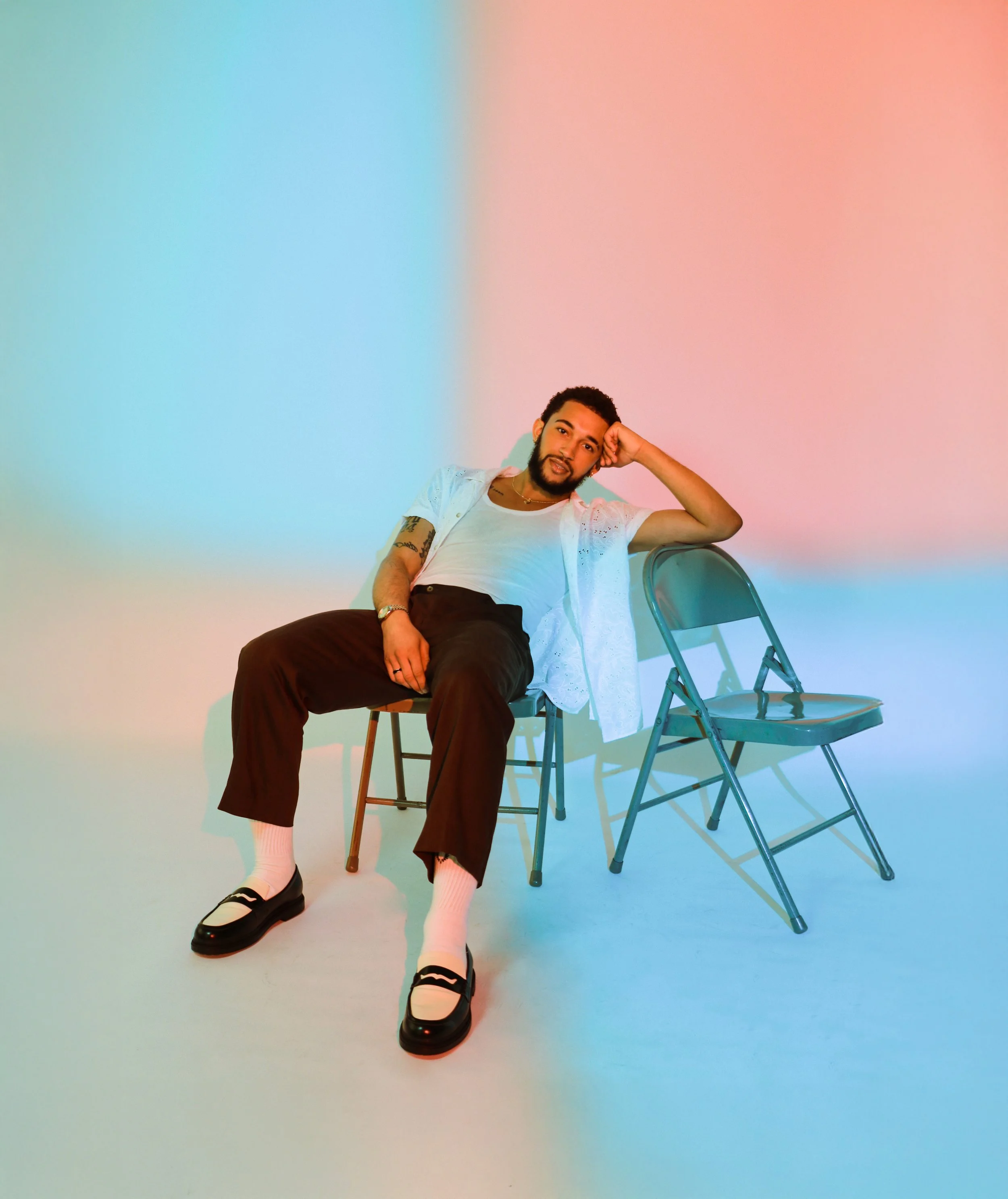A man with a beard and tattoos relaxing while sitting sideways on a metal folding chair against a pastel pink and blue background. He is wearing a white shirt, black pants, white socks, and black shoes.