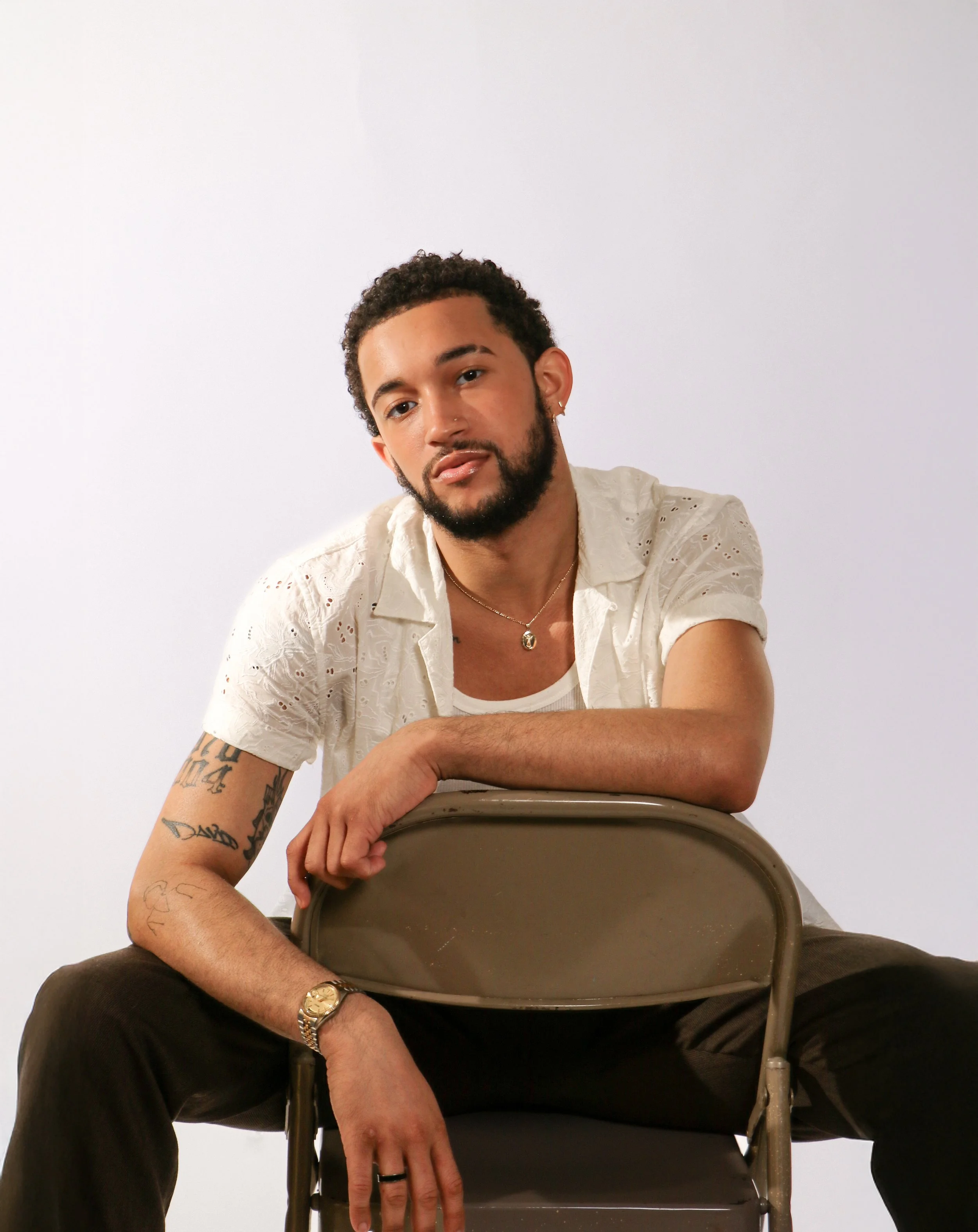 A young man with curly hair and a beard sitting on a chair, leaning forward with his arms resting on an upside down chair in front of him, wearing a white eyelet shirt, black pants, a watch, and a necklace, against a plain white background.