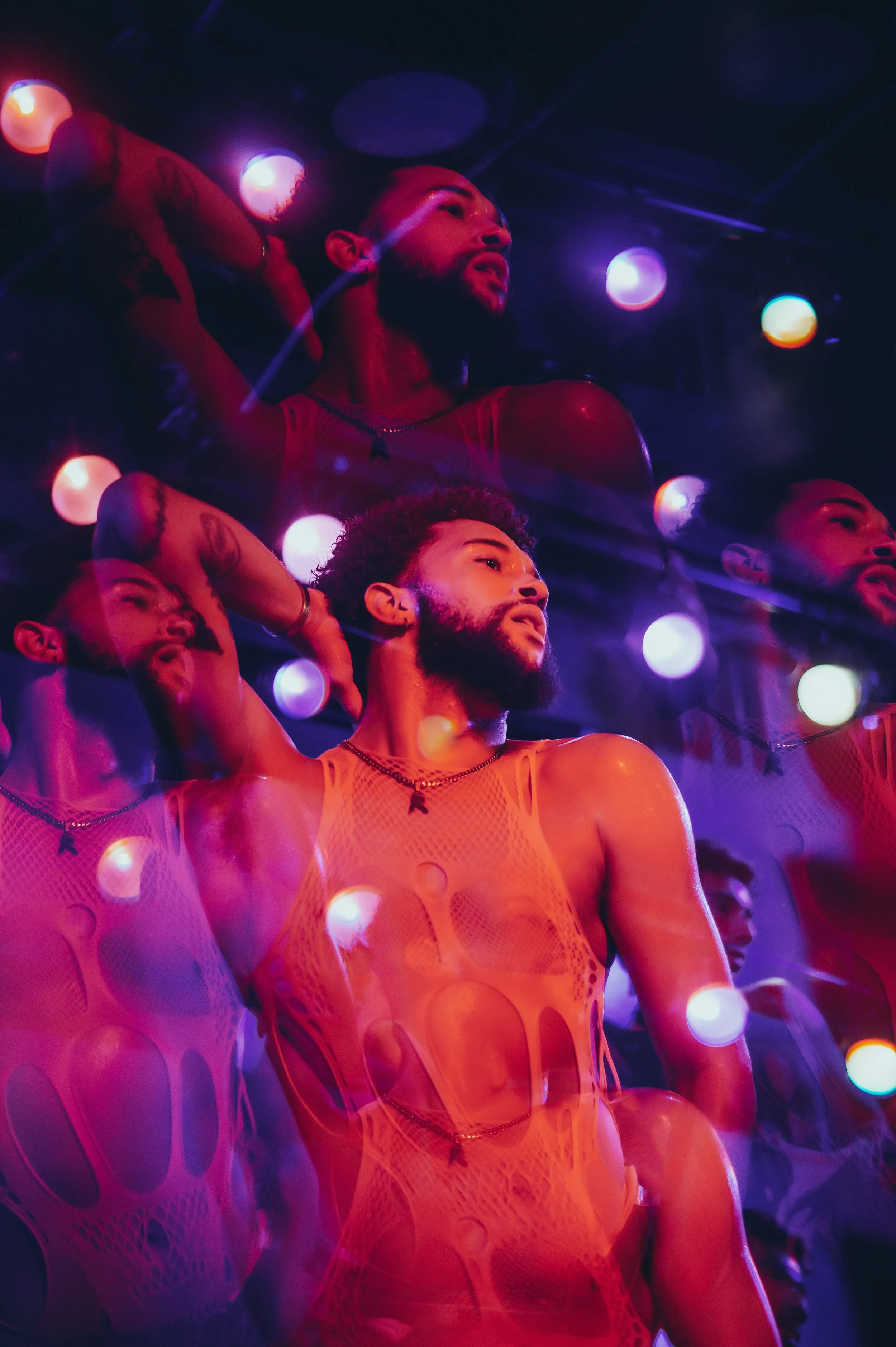 A man with a beard wearing an orange sleeveless mesh top and necklace, illuminated by colorful stage lights, with reflections of himself in the background.