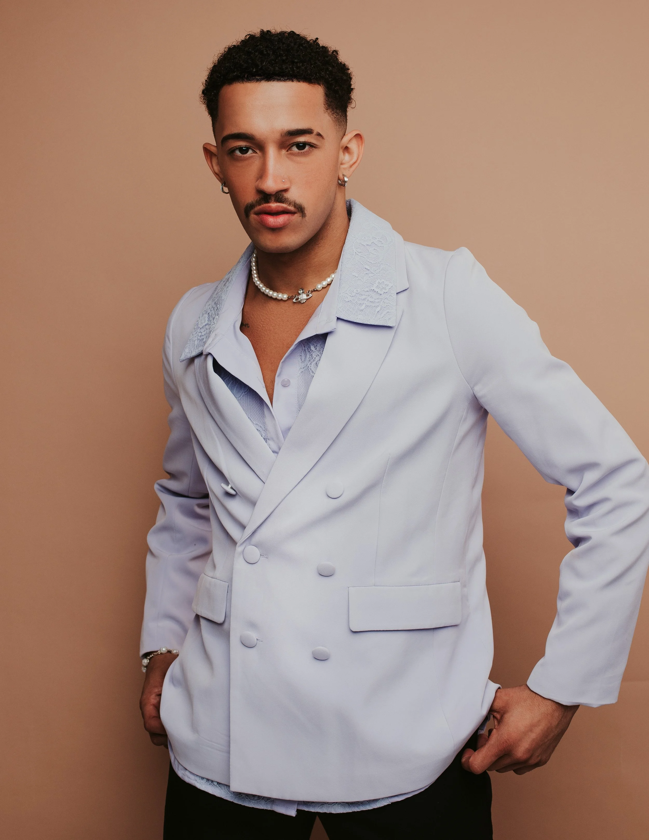 A person with short curly hair and a mustache wearing a white double-breasted blazer, gold and pearl jewelry, standing against a beige background.