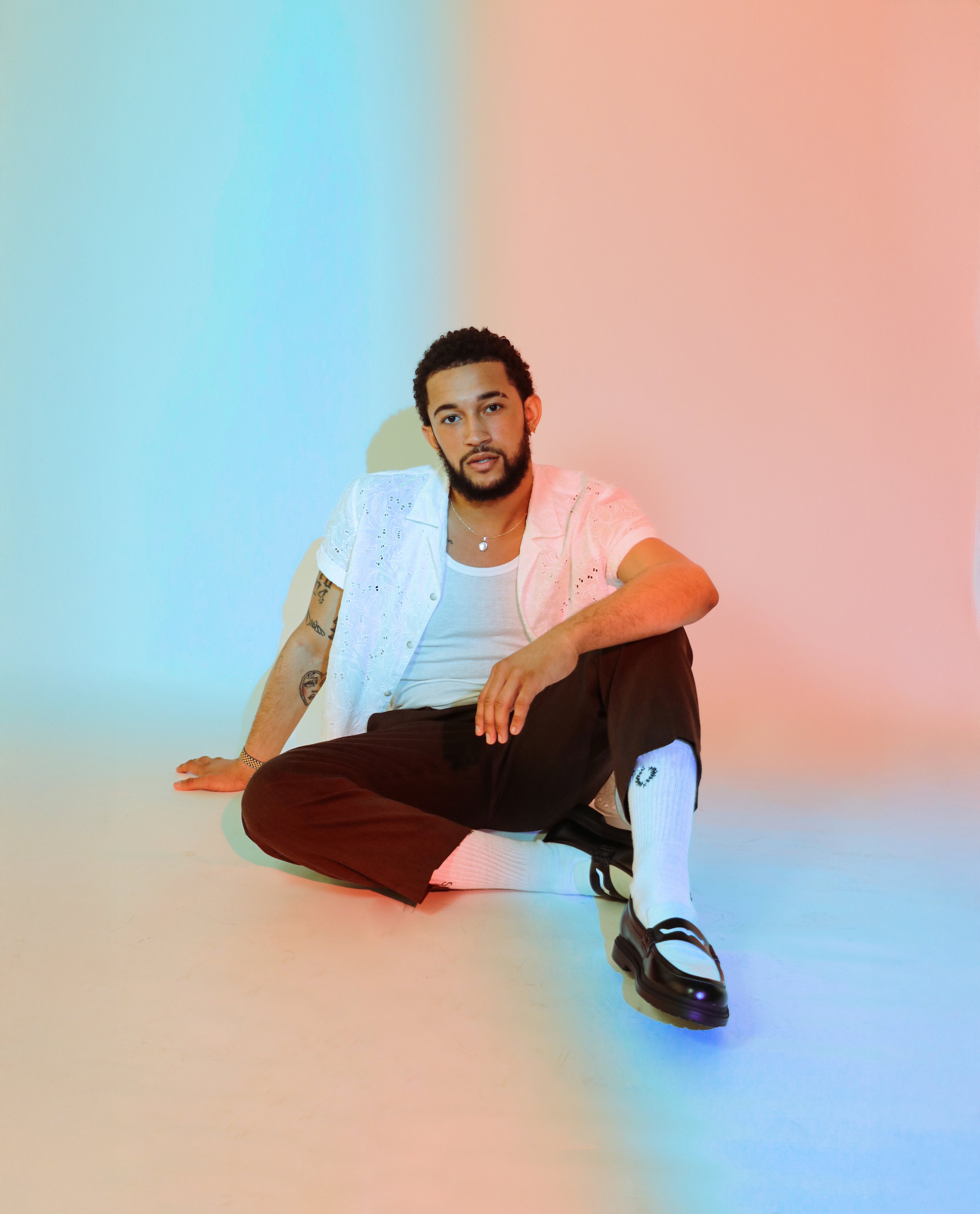 A young man with curly hair and a beard, wearing a white eyelet shirt and white socks with black sandals, poses sitting on the floor against a colorful gradient background.