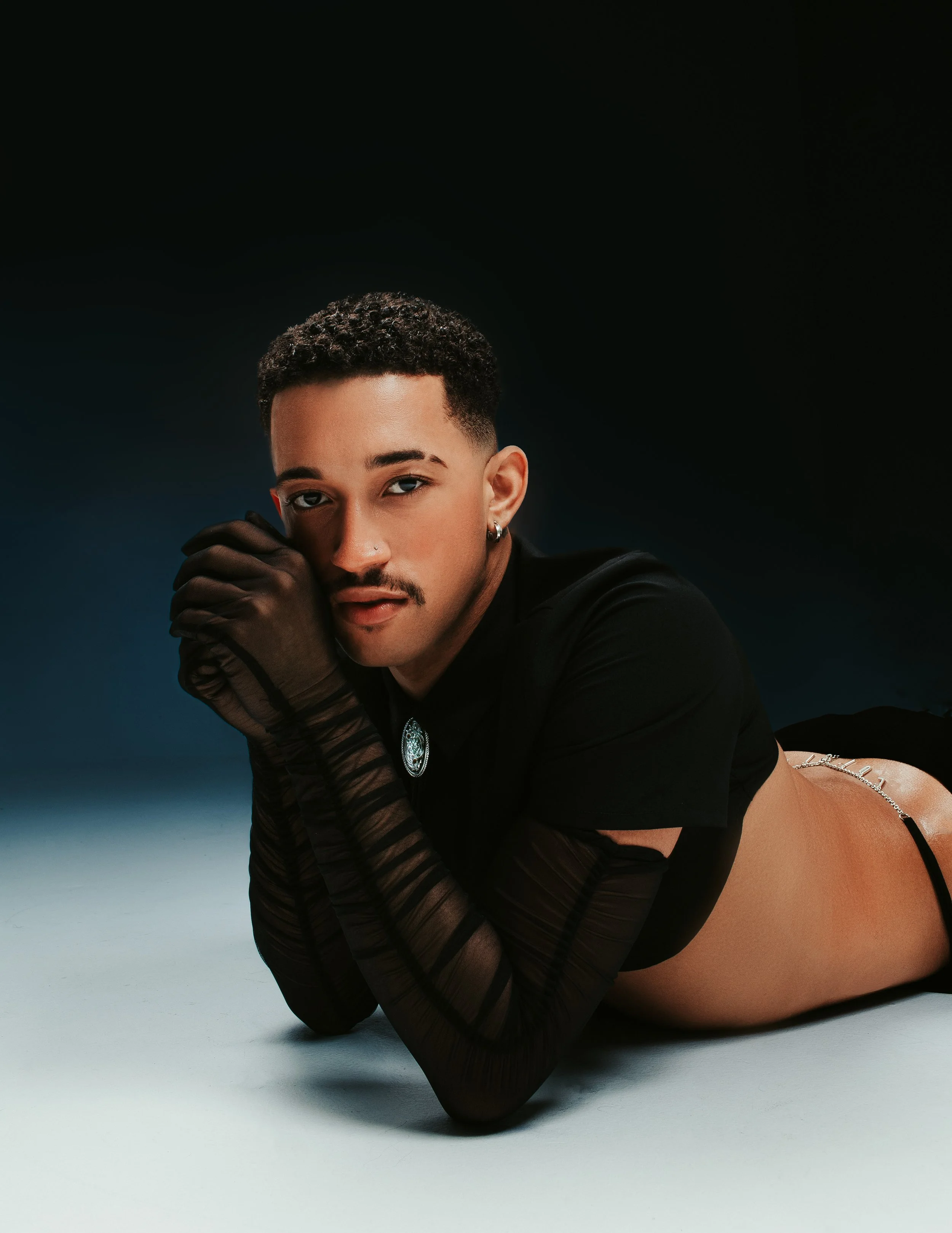 A person with short curly hair, wearing black gloves and jewelry, lying on the floor against a dark background, looking at the camera.