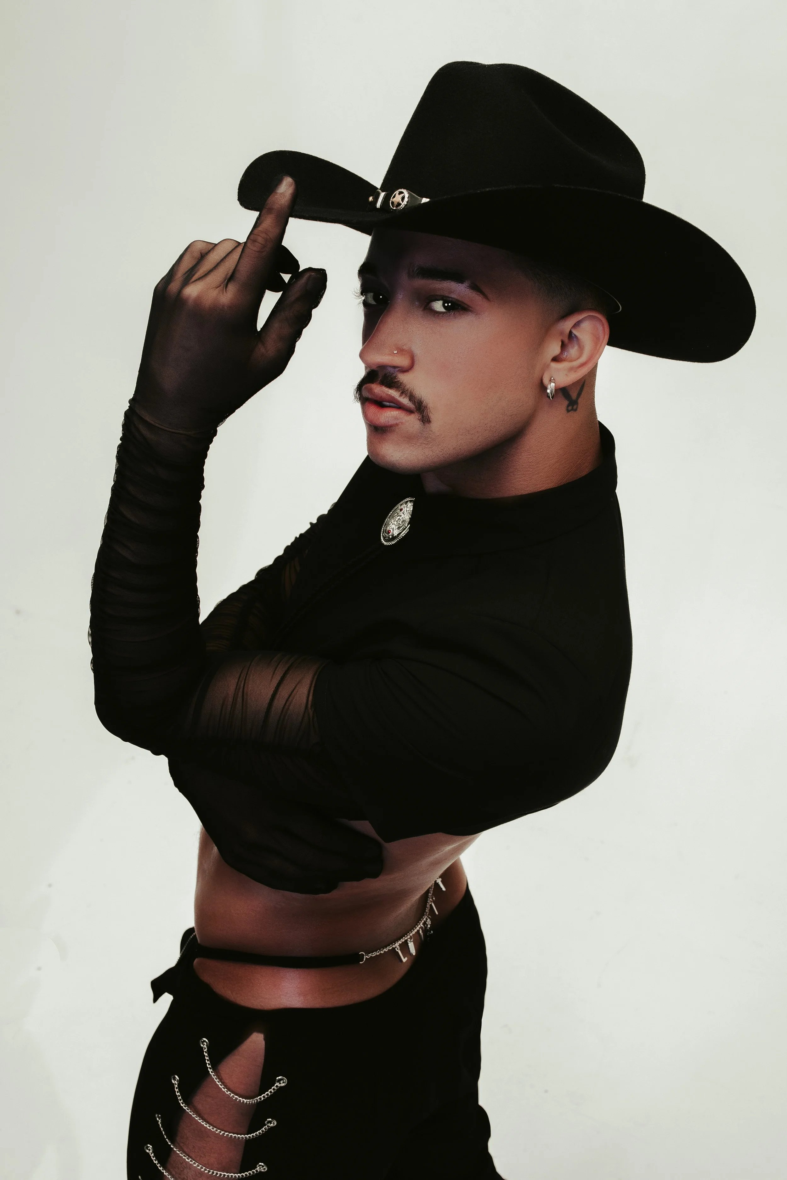 Person with short hair and tattoos wearing a wide-brimmed black hat, black sheer top, and jewelry, tilting the hat with their hand, against a plain background.