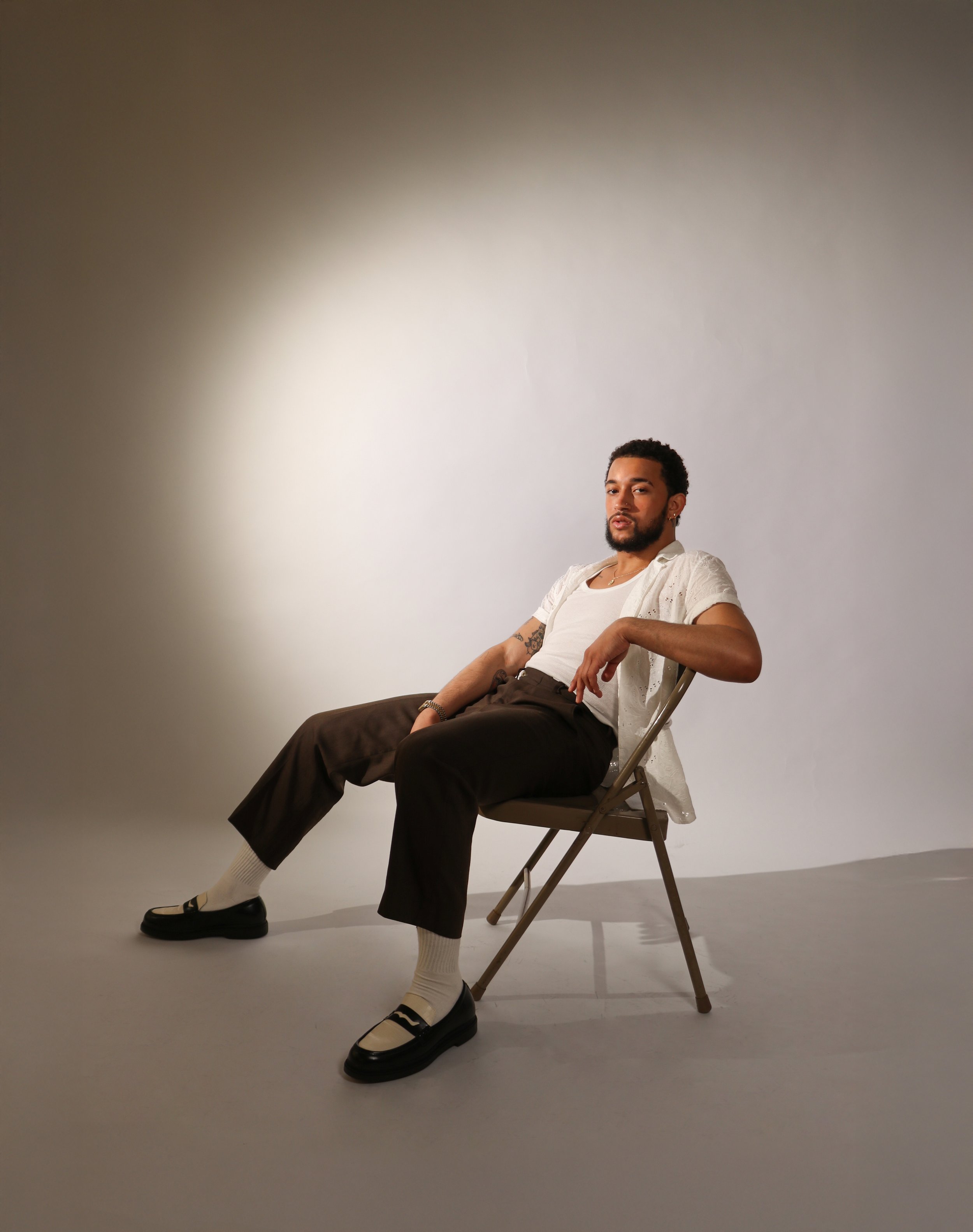 A young man with dark curly hair, a beard, and earrings, sitting on a chair with his legs crossed. He is wearing a white shirt with short sleeves, dark pants, cream socks, and black shoes with white accents. The background is plain and neutral with a