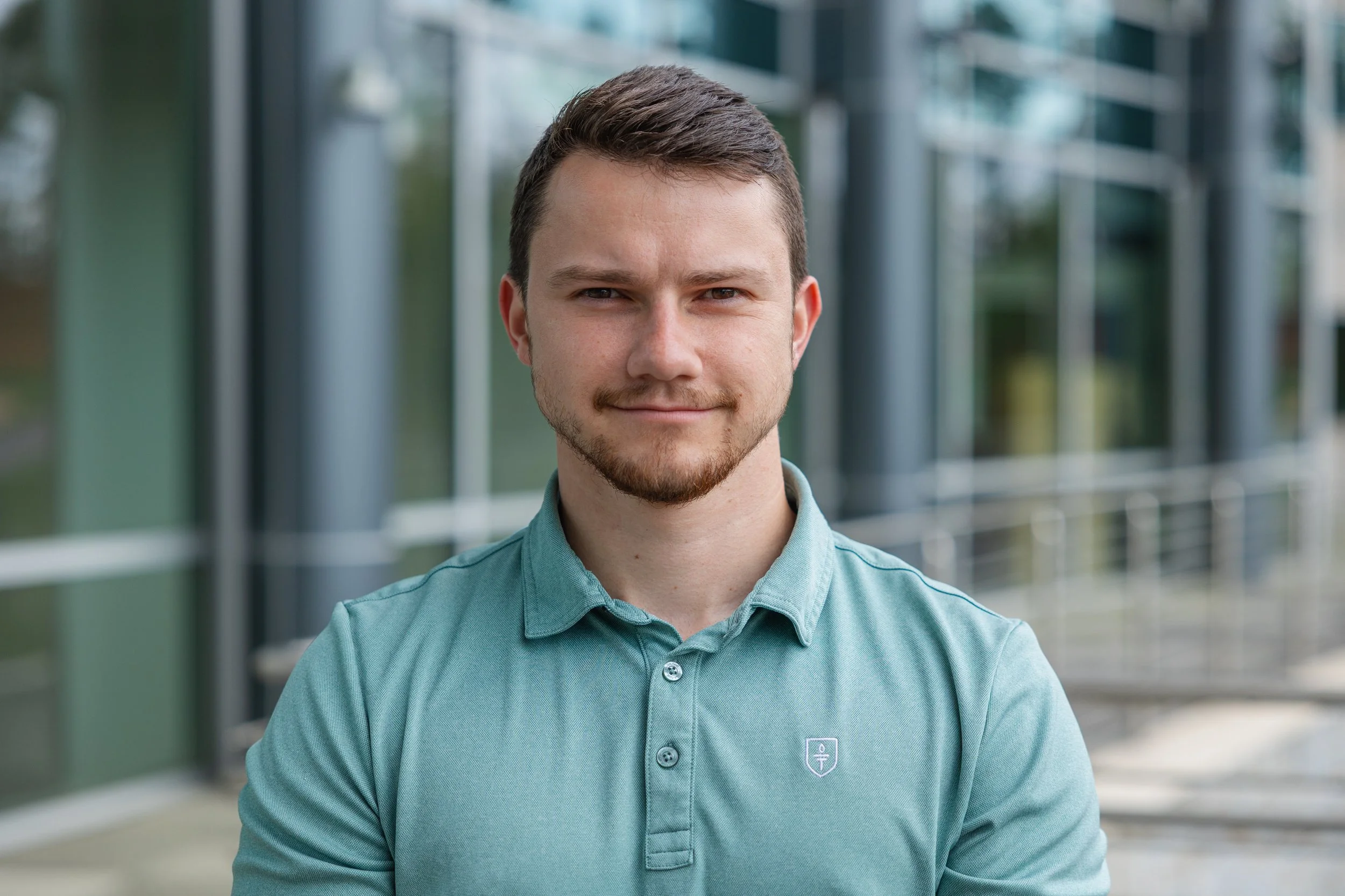 A young man with short brown hair and a beard, wearing a teal button-up shirt with a small logo on the chest, smiling slightly, standing outside in front of a modern glass building.