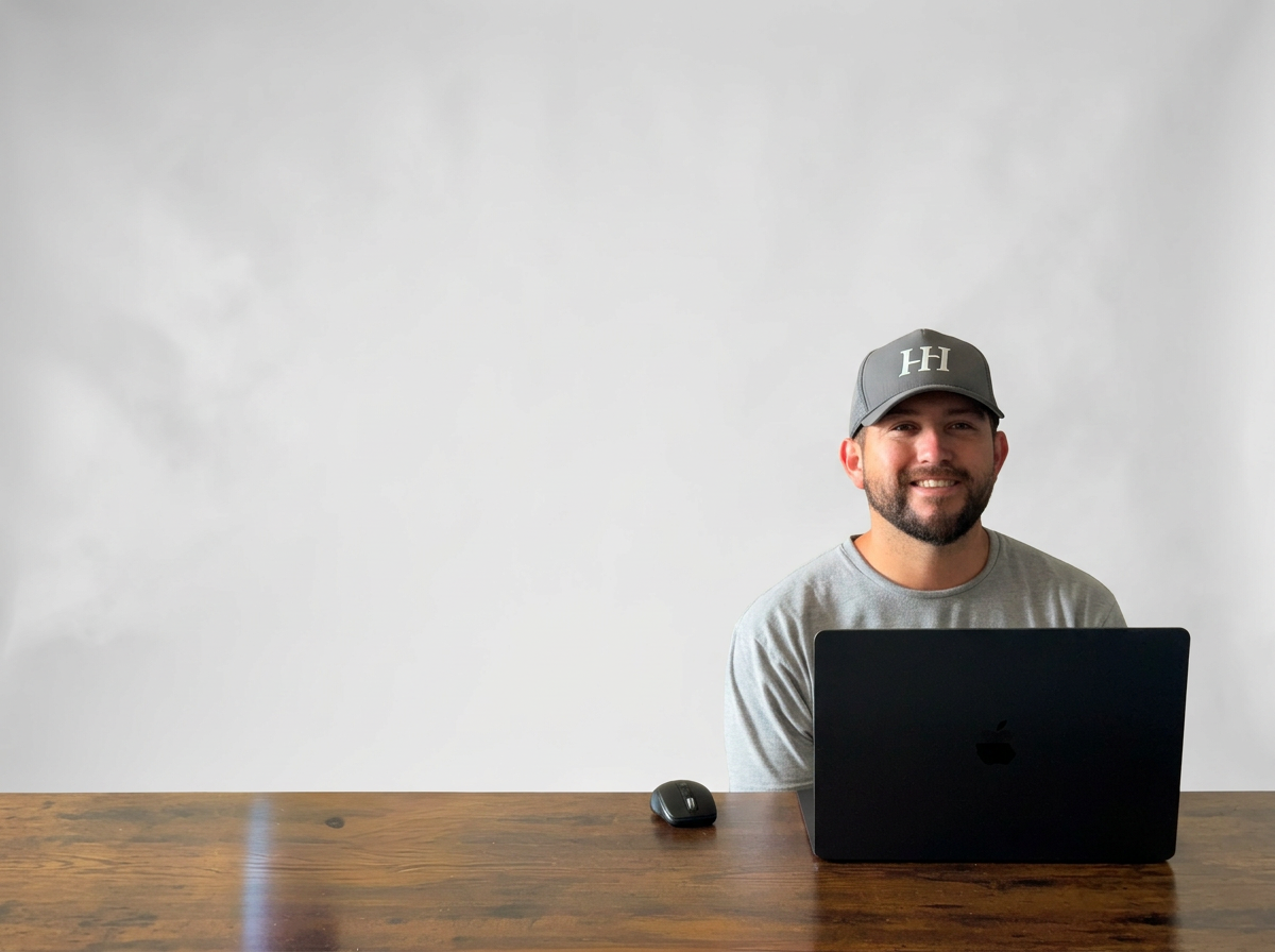 A man with a beard wearing a gray cap and gray t-shirt sitting at a wooden desk with a black laptop and mouse, smiling at the camera, in front of a plain white wall.