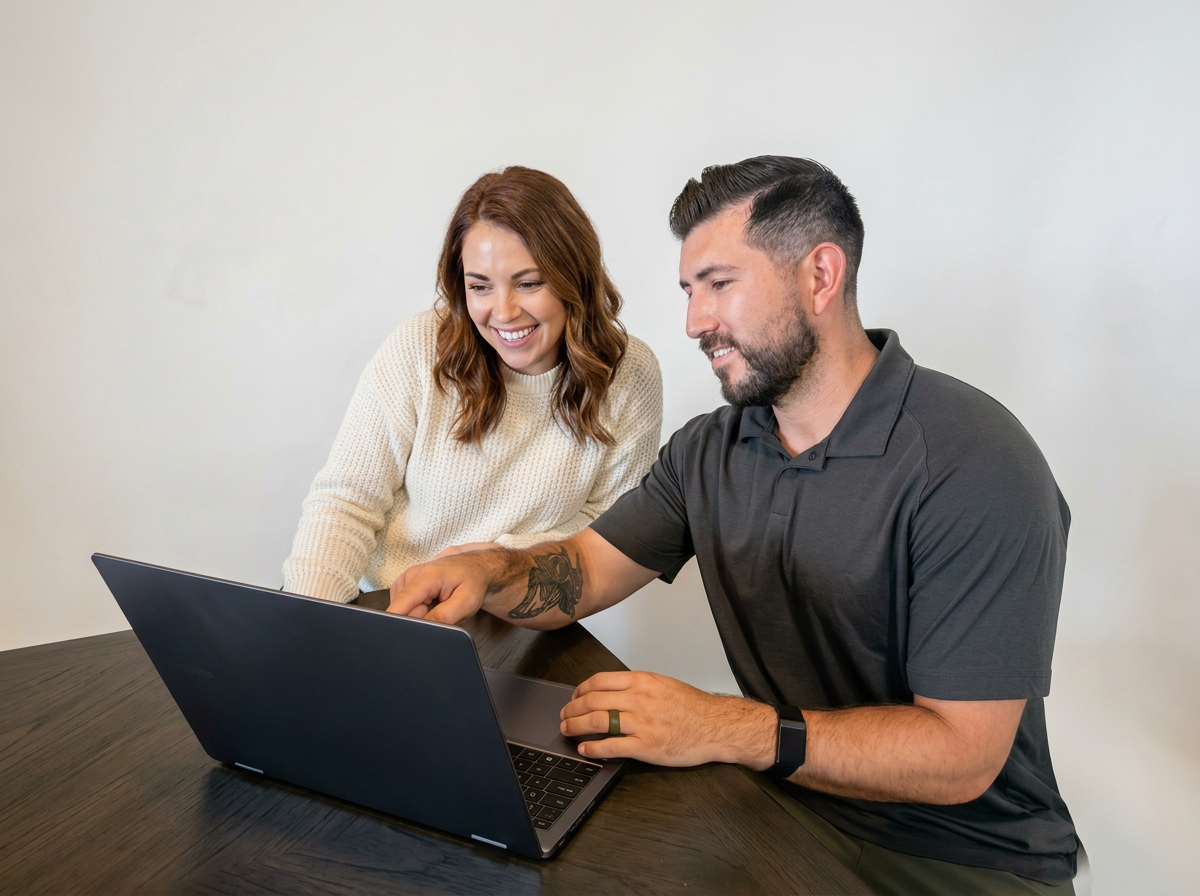 A woman and a man are sitting at a table looking at a laptop screen and smiling.
