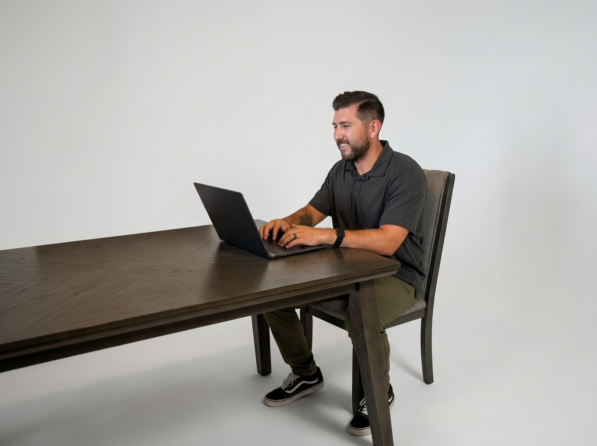 A man with dark hair and a beard sitting at a dark wooden desk, working on a laptop, against a plain white background.