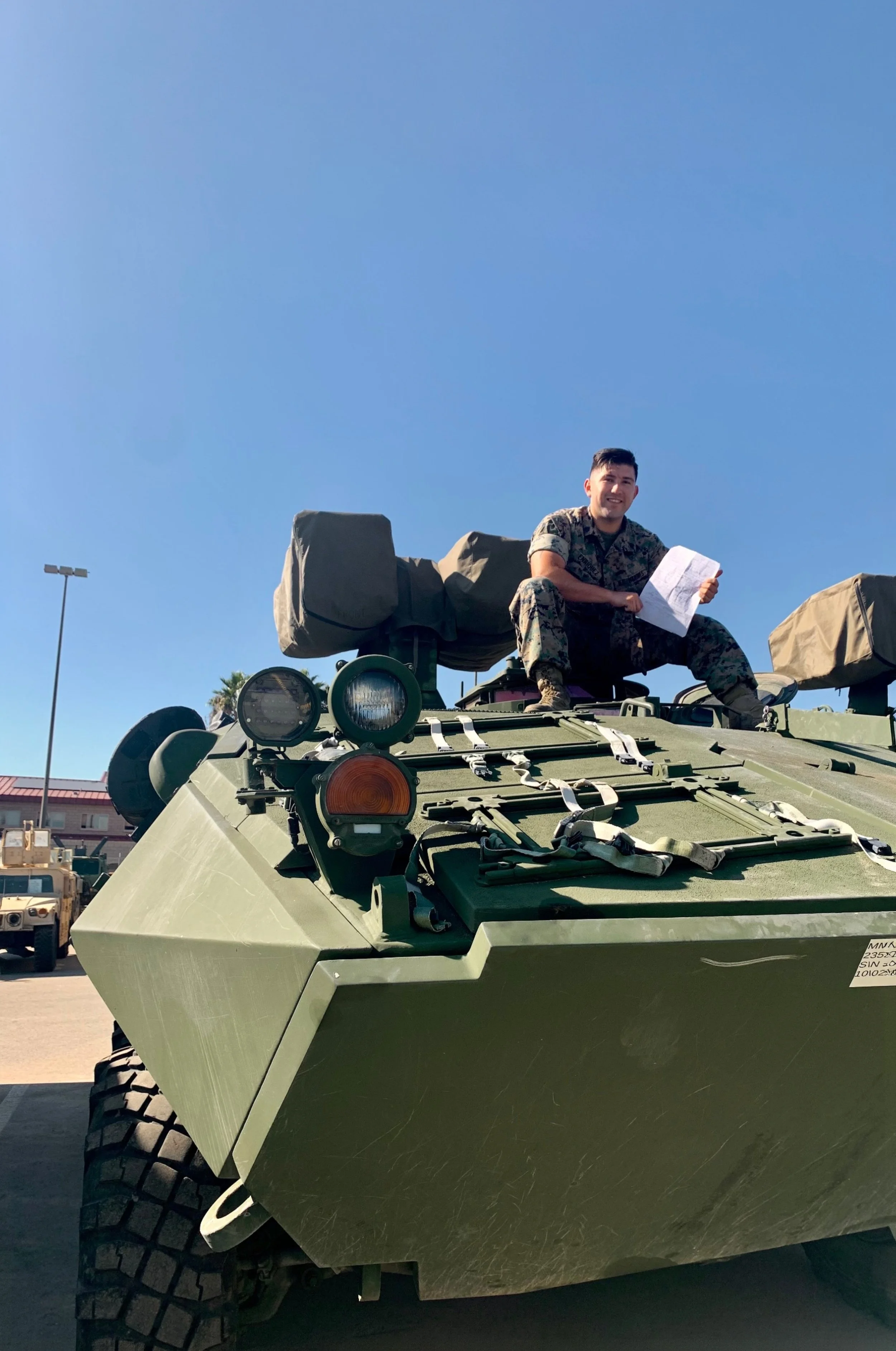 A soldier in camouflage uniform sitting on top of a green military tank holding a piece of paper, with clear blue sky in the background.