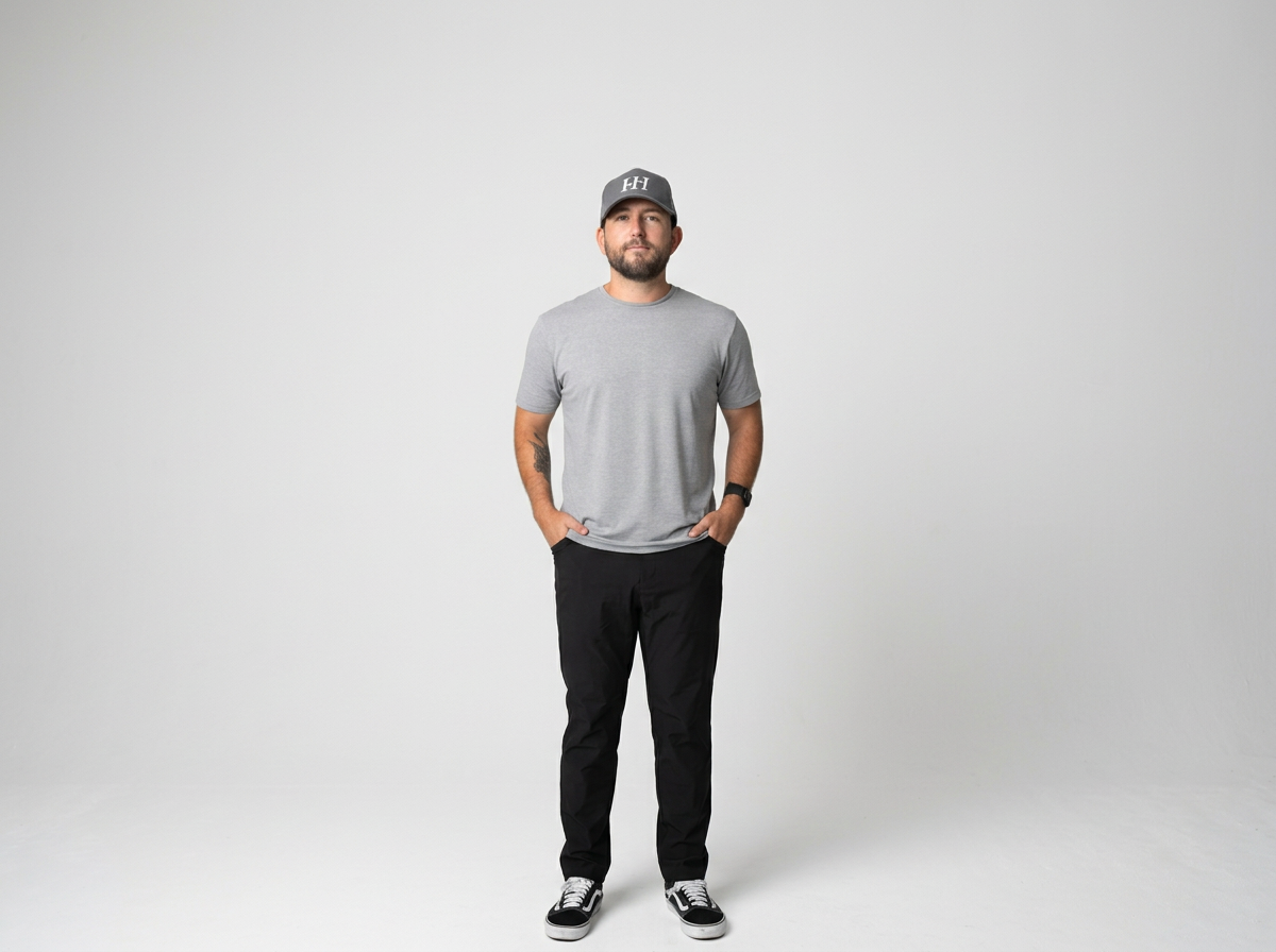 Man wearing a grey T-shirt, black pants, sneakers, and a baseball cap standing against a plain white background.