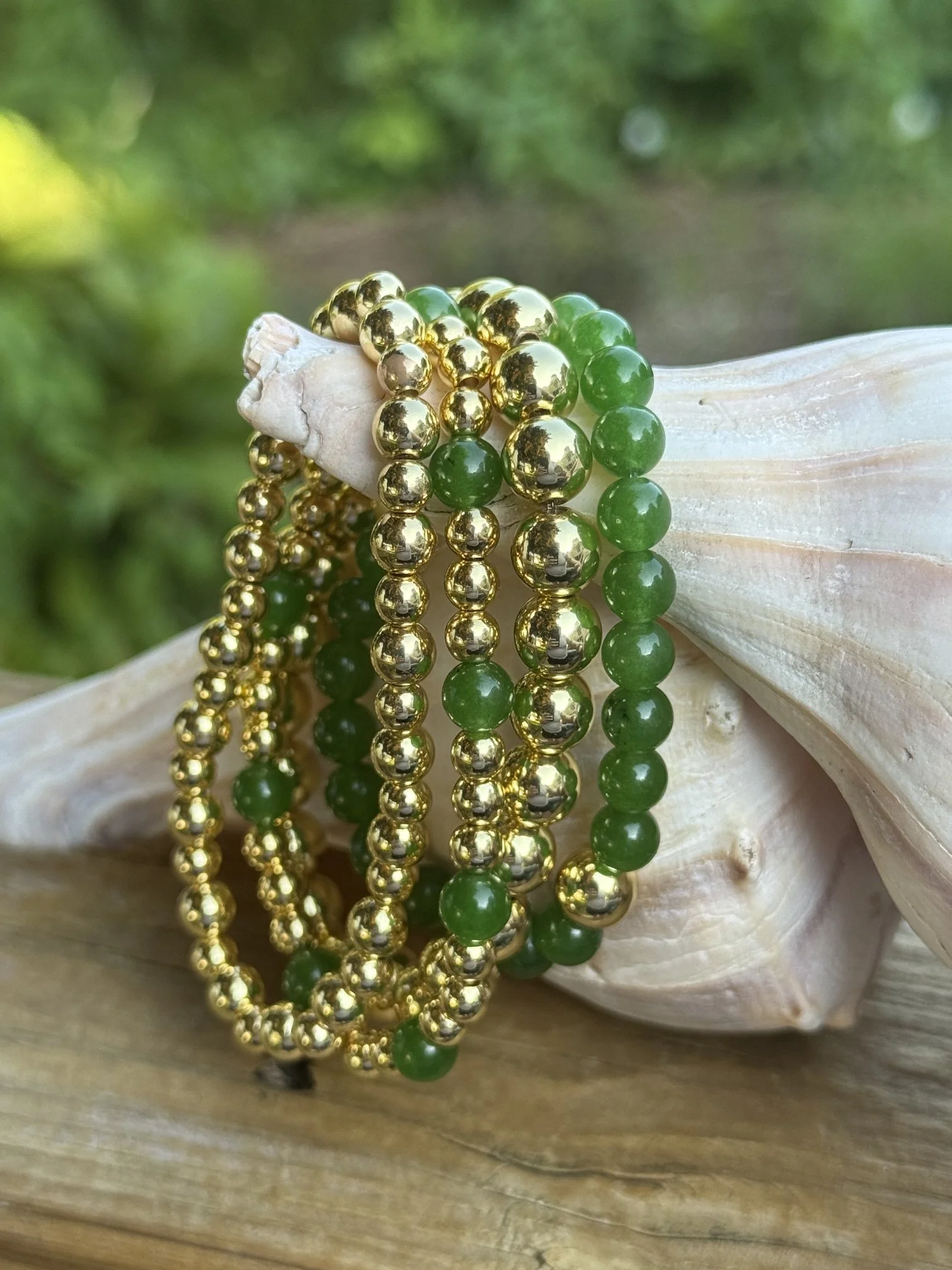 Multiple strands of green and gold beads draped over a white seashell, with a blurred green background.