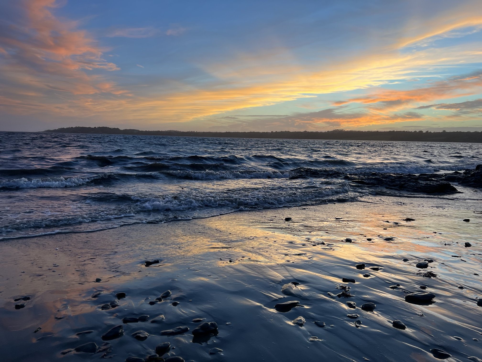A beach scene during sunset with the sky painted in shades of pink, orange, and blue. The ocean has gentle waves reaching the sandy shore, which is scattered with small rocks and pebbles.