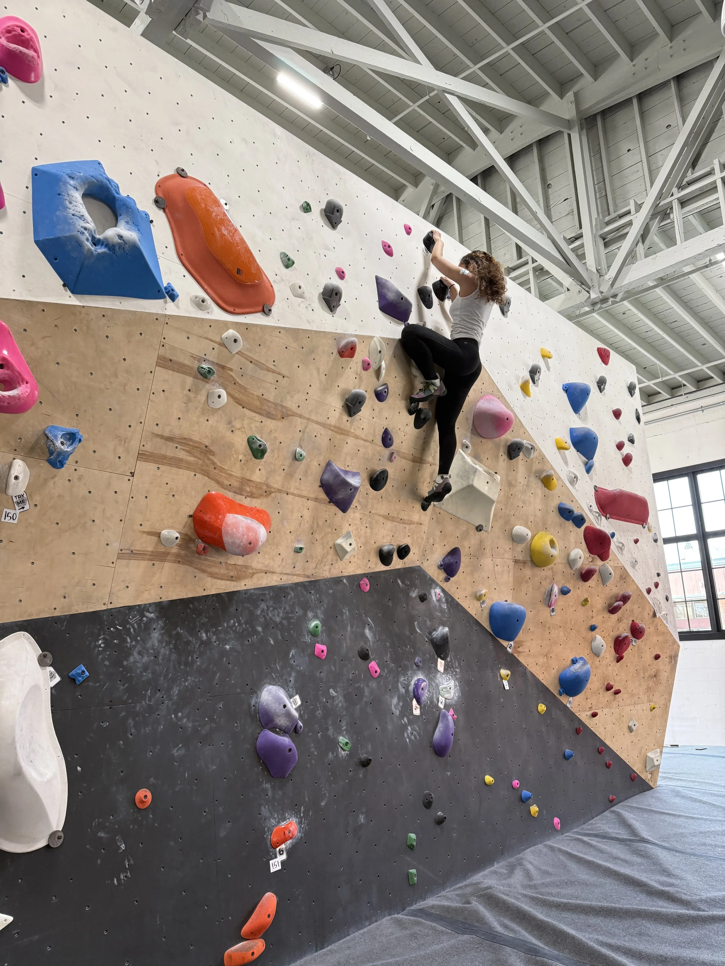 Indoor bouldering gym with a woman climbing on a wall with colorful holds.