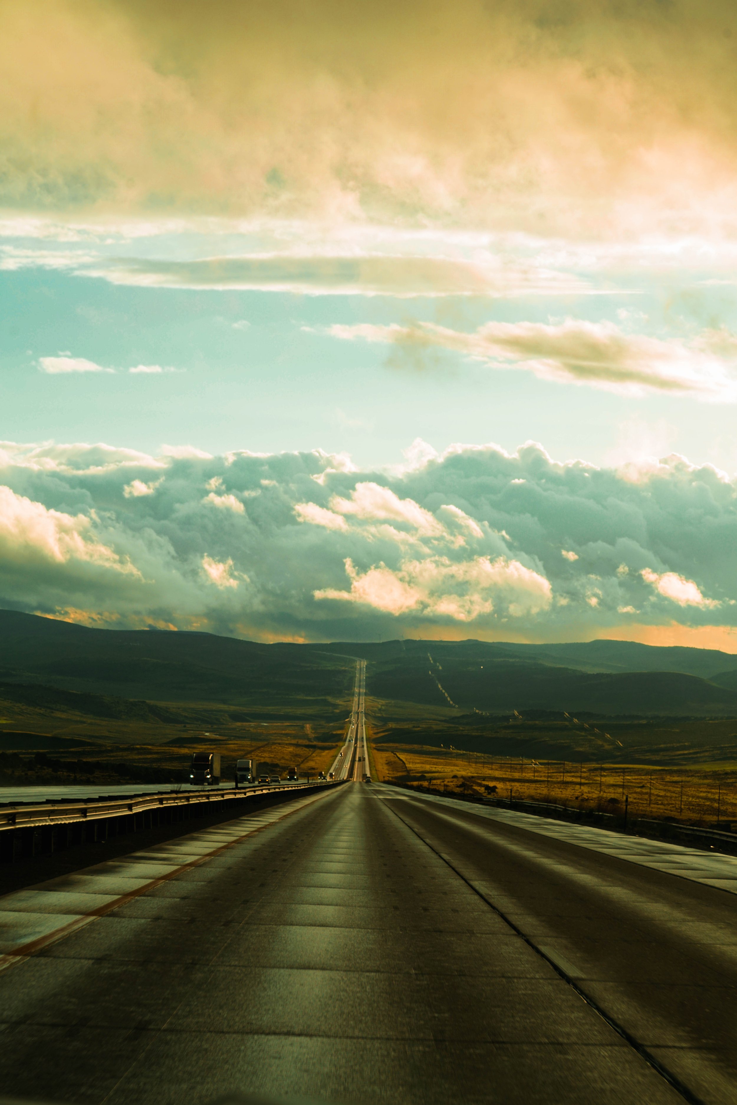 Long straight highway stretching into the horizon with cars driving on it. The landscape is hilly with green and brown fields and distant mountains. The sky has a mixture of bright clouds and darker clouds with sunlight breaking through.