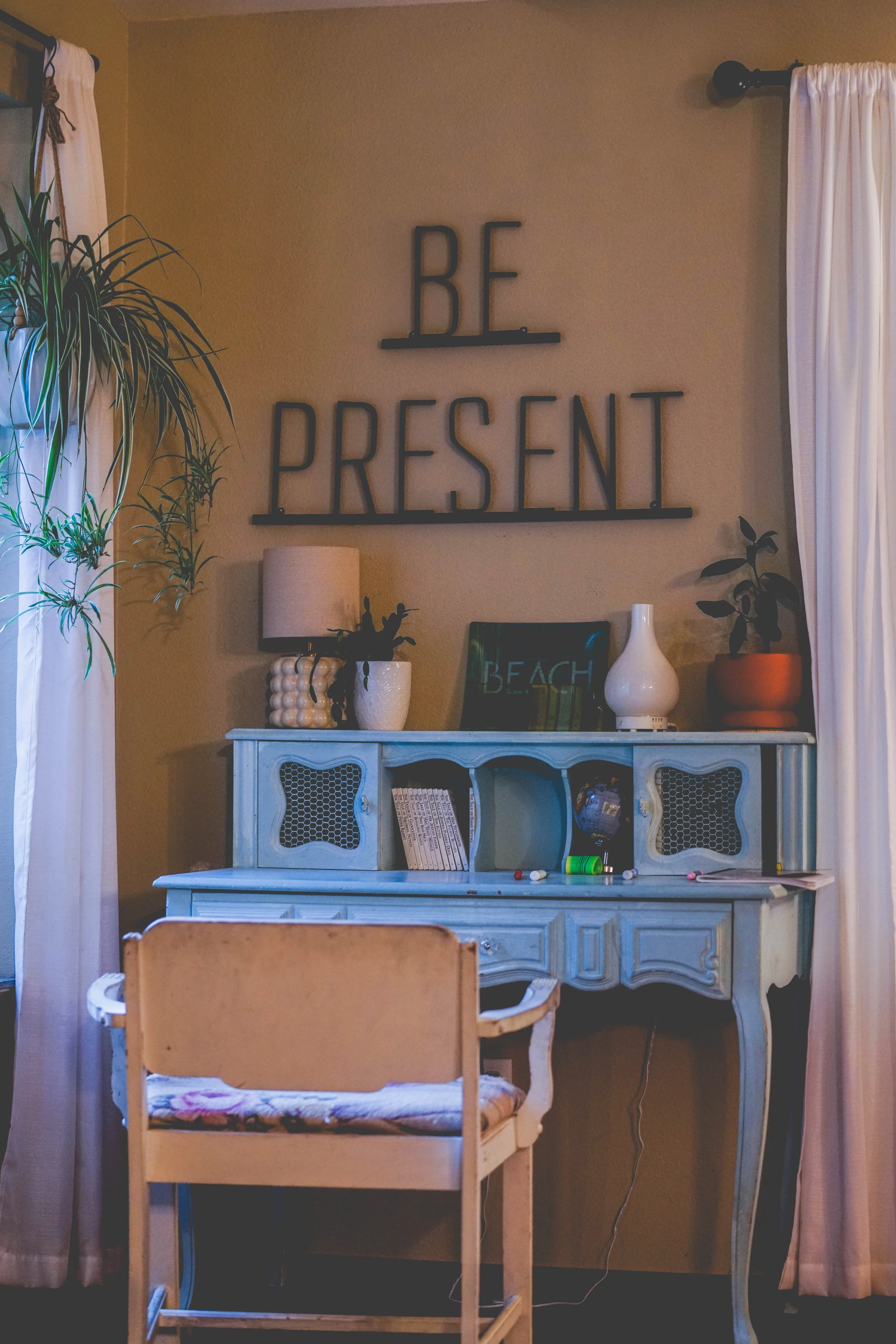 A cozy living room corner with a decorated light blue vintage hutch holding various decorative items, a small table, and an armchair. The wall above the hutch features a black metal sign that reads 'BE PRESENT'. There are green plants on either side of the setup, white curtains, and a lamp on the shelf to the left.