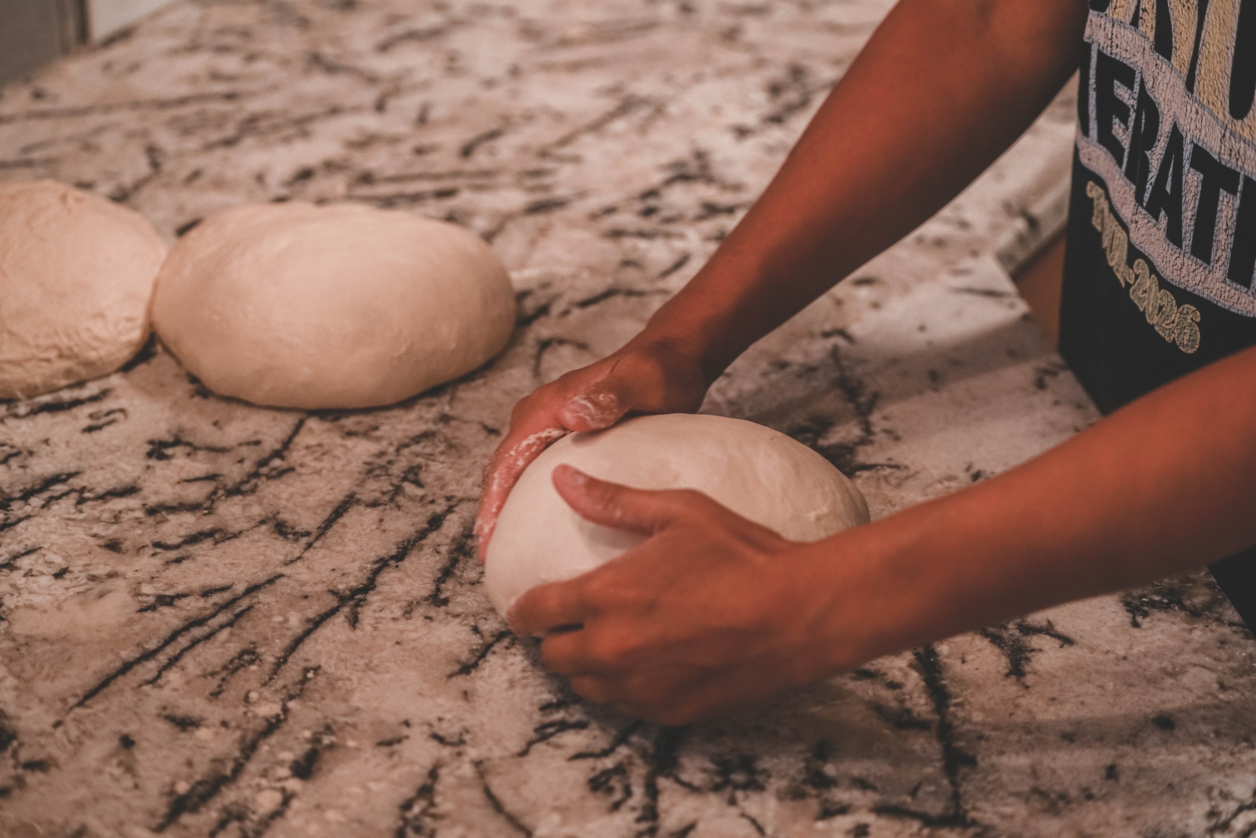 A person kneading dough on a marble countertop with their hands, wearing a shirt with colorful text.