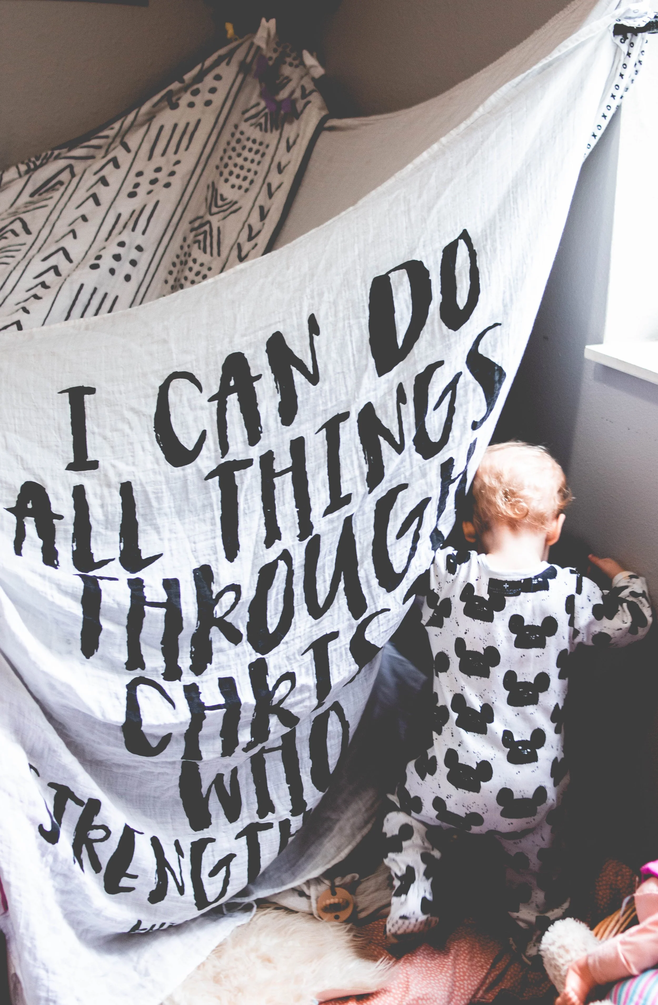 Children playing under a blanket with alphabet and skeleton patterns, with a banner reading 'I CAN DO ALL THINGS' partially visible.
