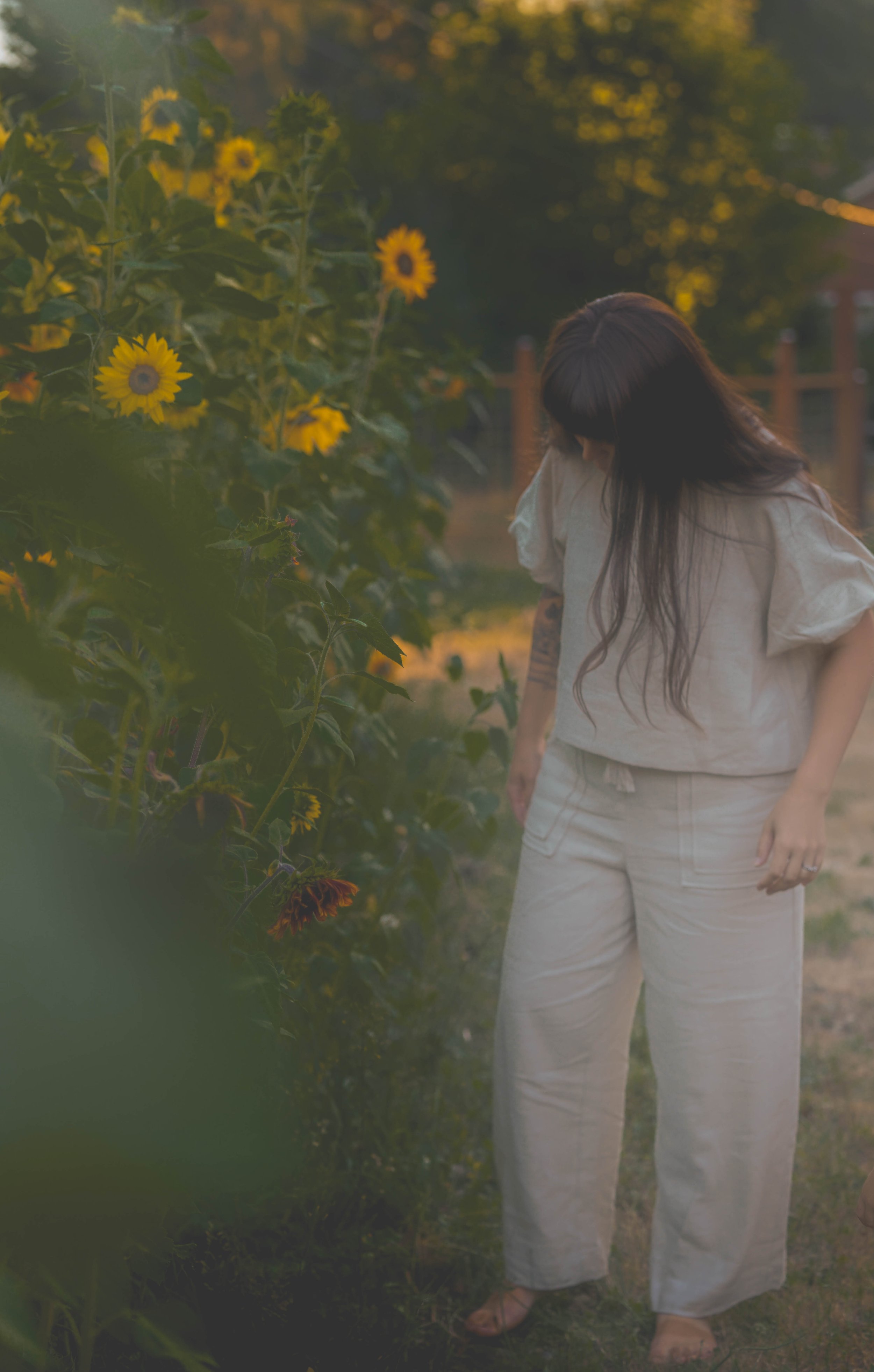Woman with long dark hair wearing beige clothes standing in a garden during sunset, surrounded by yellow flowers and greenery.