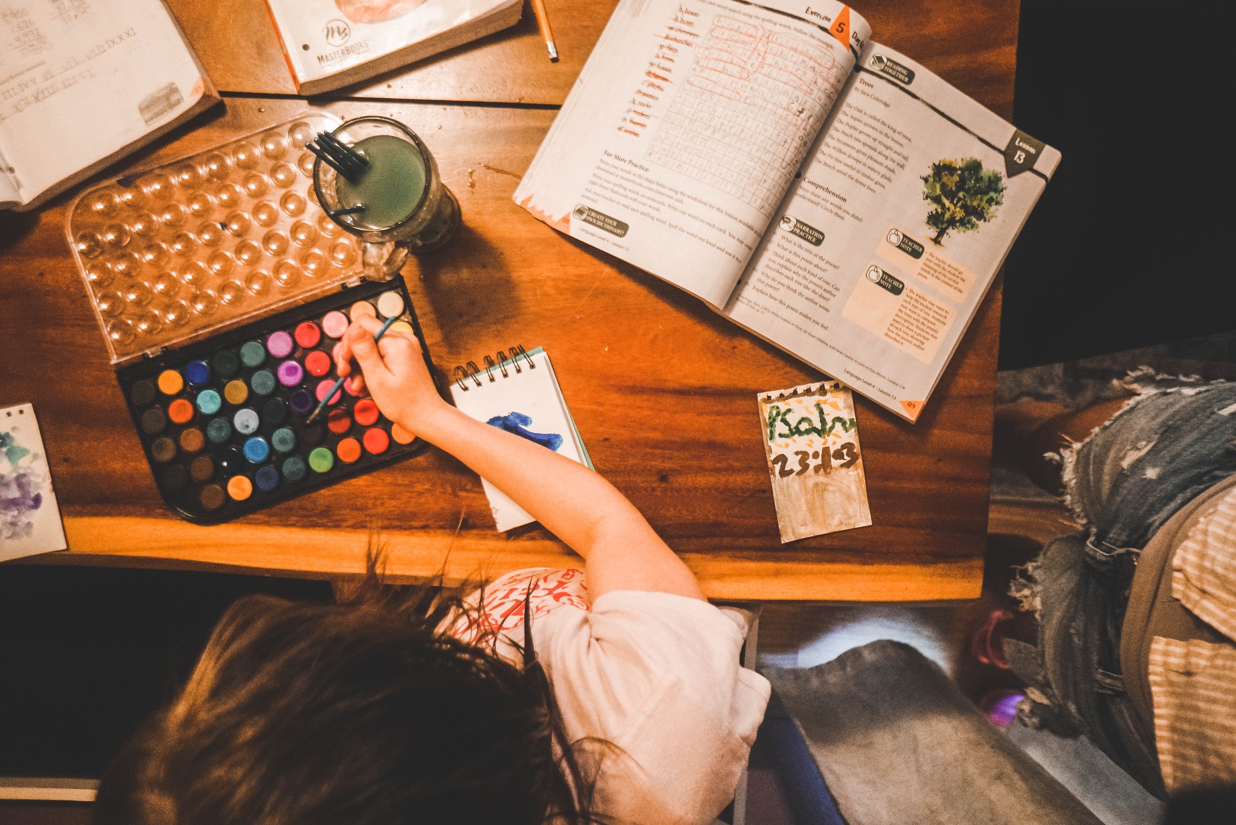 Top-down view of a wooden table with open books, a paint set, a glass with a drink and some DIY materials, a person painting with a small brush, and various papers and notebooks scattered around.