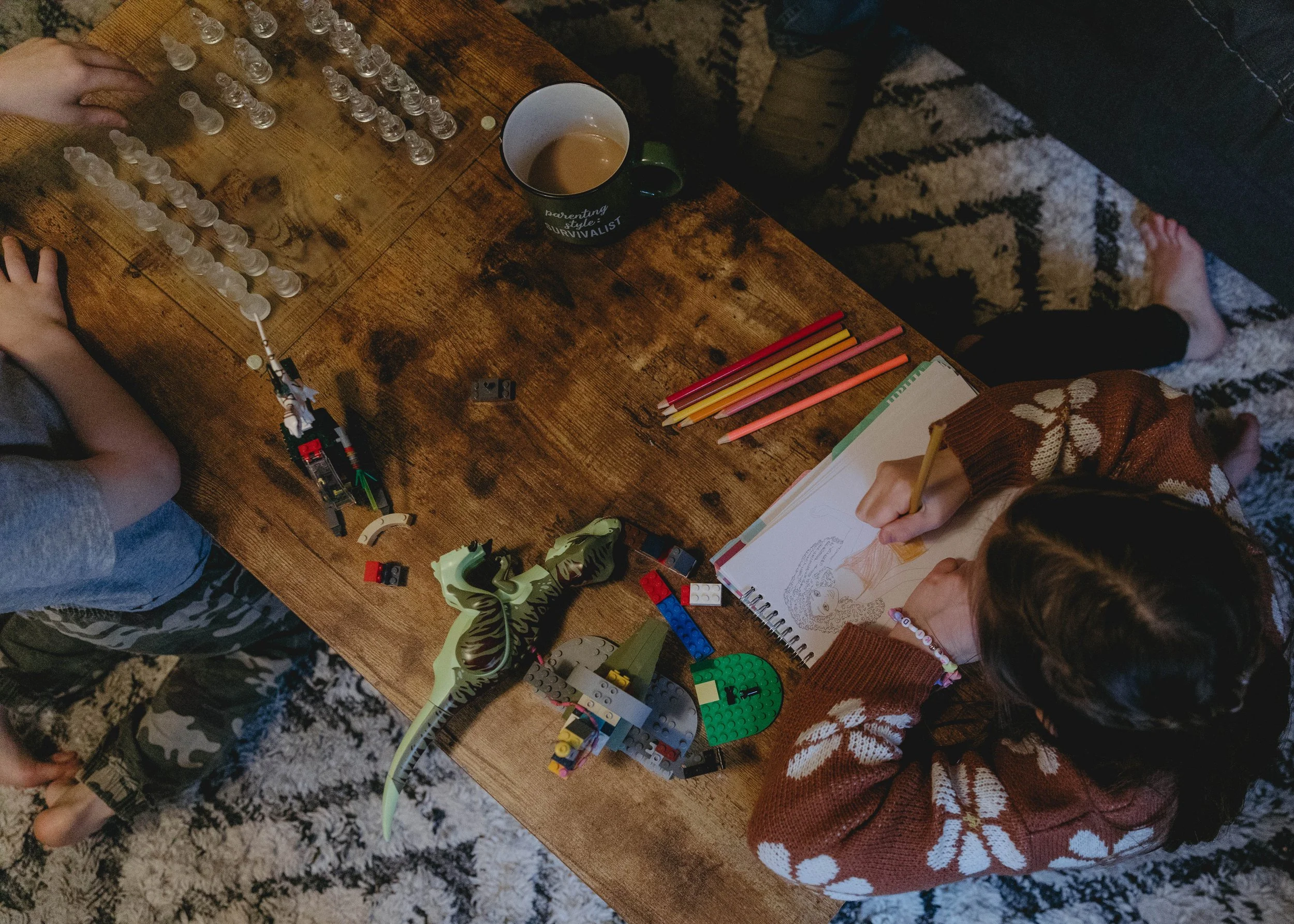 Children playing with Lego blocks on a wooden table, one is drawing in a notebook, a mug with coffee, and a green dinosaur toy are also on the table.