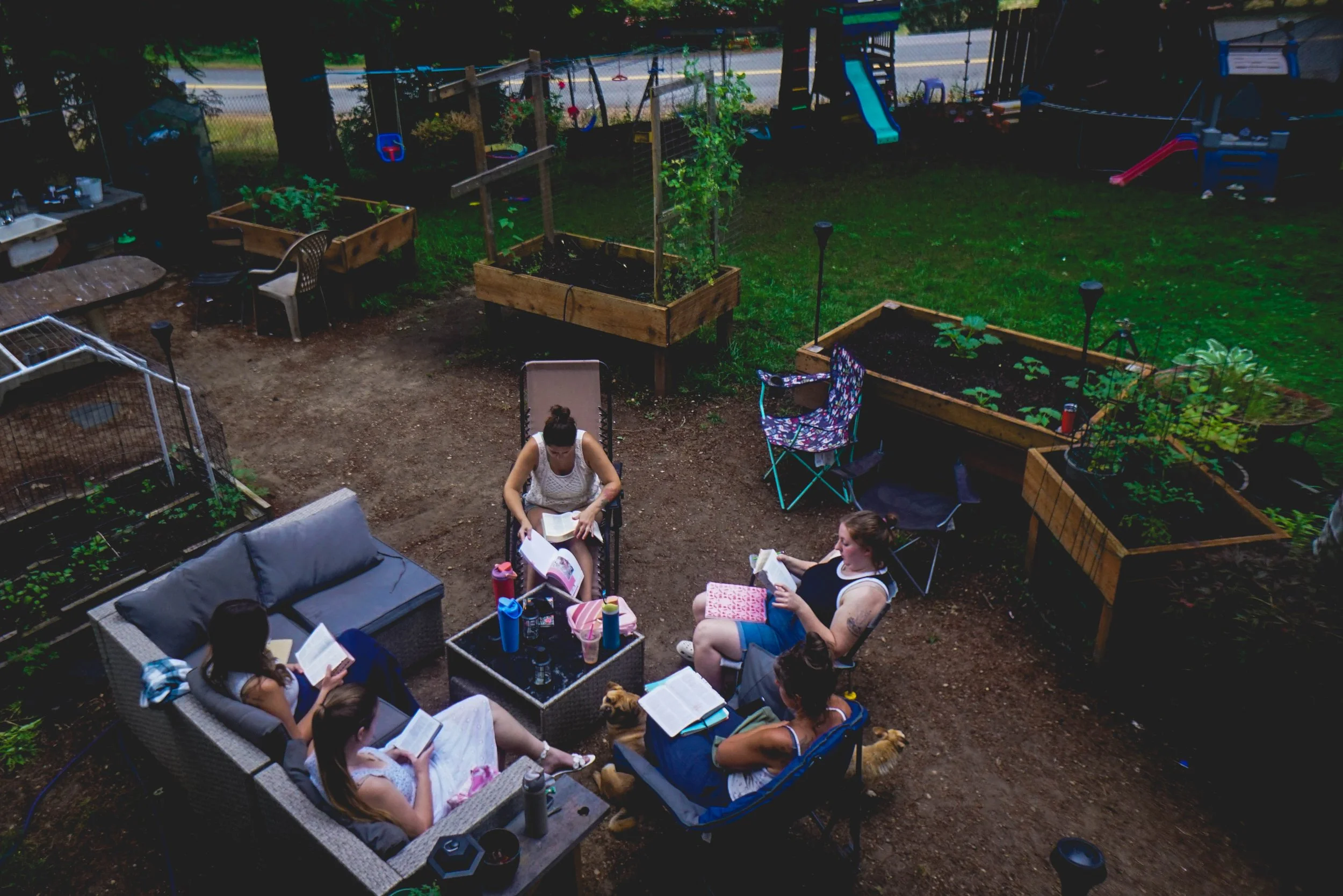 People relaxing in a backyard garden, sitting on outdoor furniture and reading books, with plants and children's play equipment in the background.