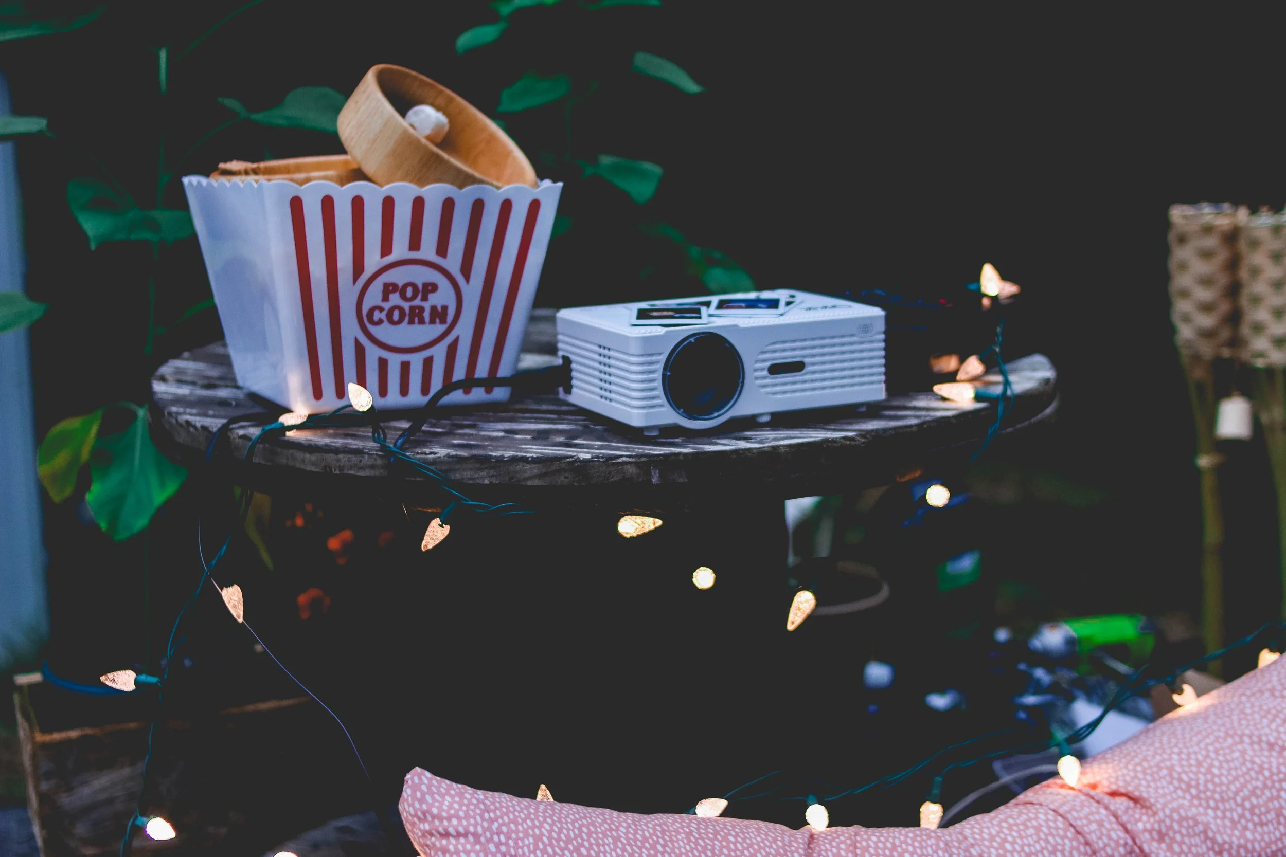 A wooden bowl with a popcorn container, a retro camera, and string lights on a wooden surface with green leafy plants in the background.
