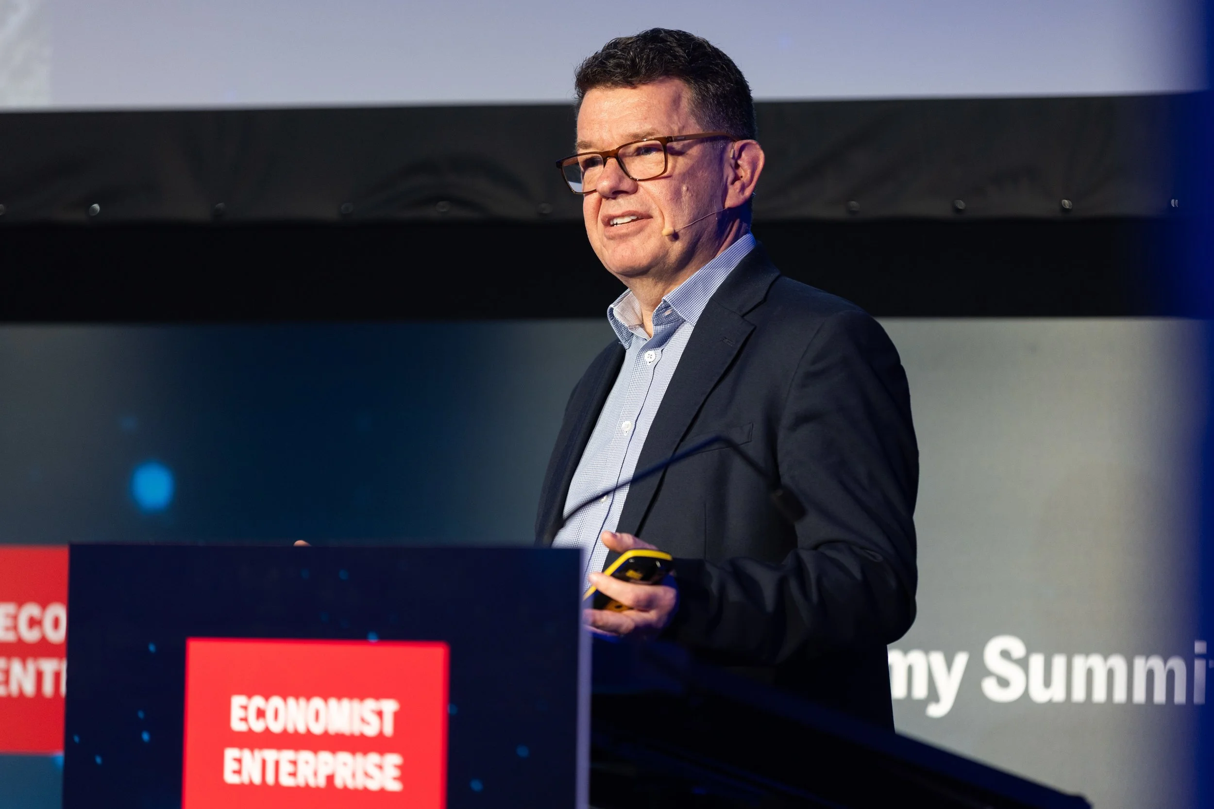 A man with dark hair, glasses, and a suit presenting at a conference. He is holding a yellow object in his right hand. The background includes a screen with the partially visible text 'my Sum...' and a red banner with the word 'ECONOMIST' repeated.