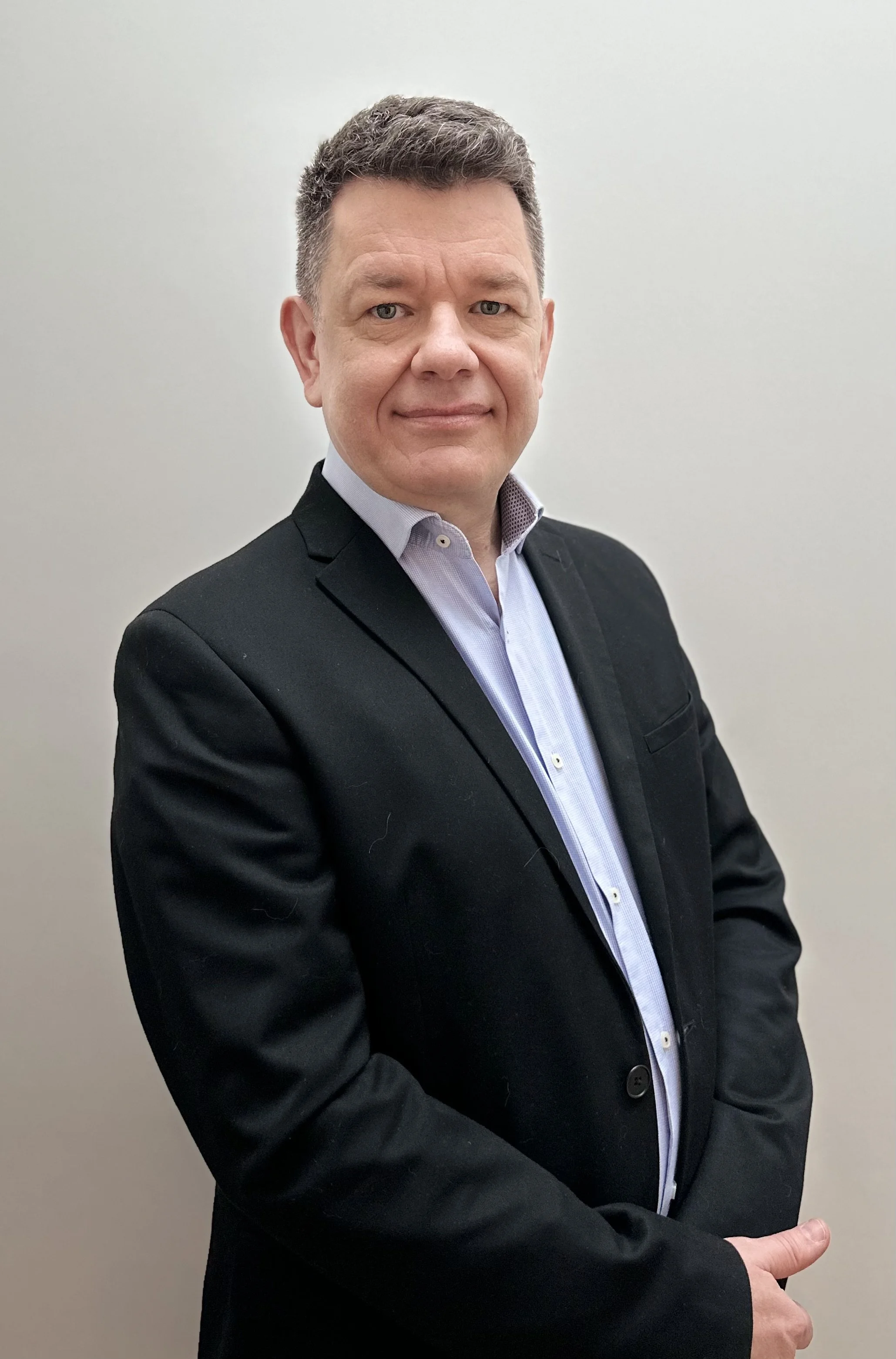 A middle-aged man with short, dark hair, dressed in a black suit and light blue shirt, standing against a plain white wall, looking at the camera with a neutral expression.