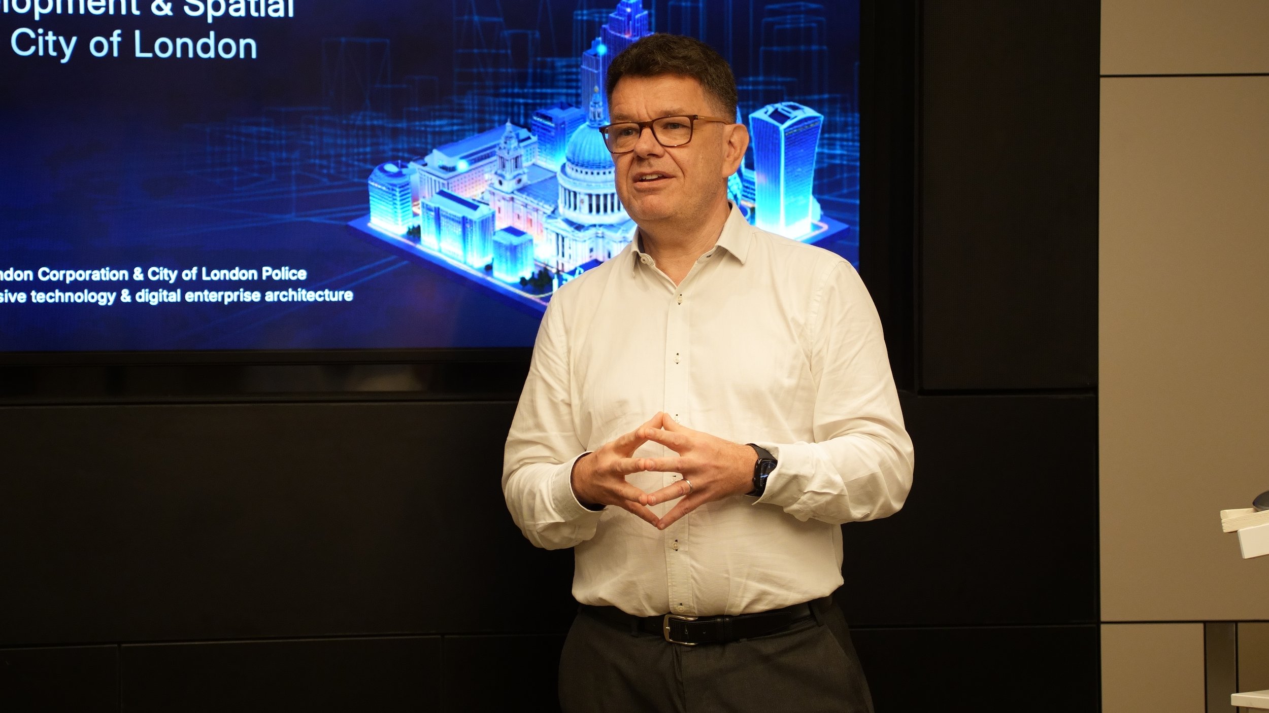 A man wearing glasses and a white shirt is speaking in front of a screen that displays a digital rendering of London's cityscape, with text mentioning 'City of London' and 'London Corporation & City of London Police'.