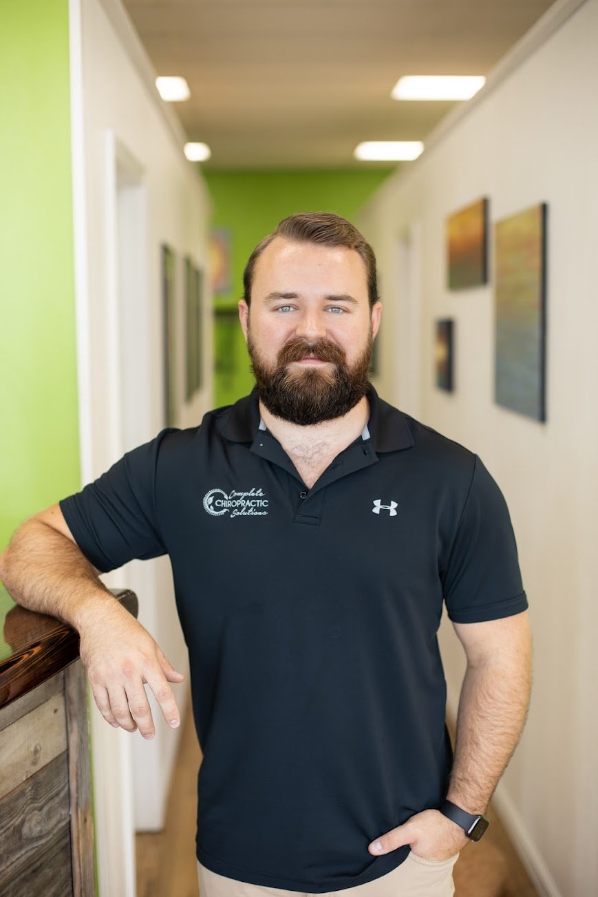 A man with a beard and blue eyes standing in a hallway with green and white walls, wearing a black polo shirt with logo and text, and smiling at the camera.