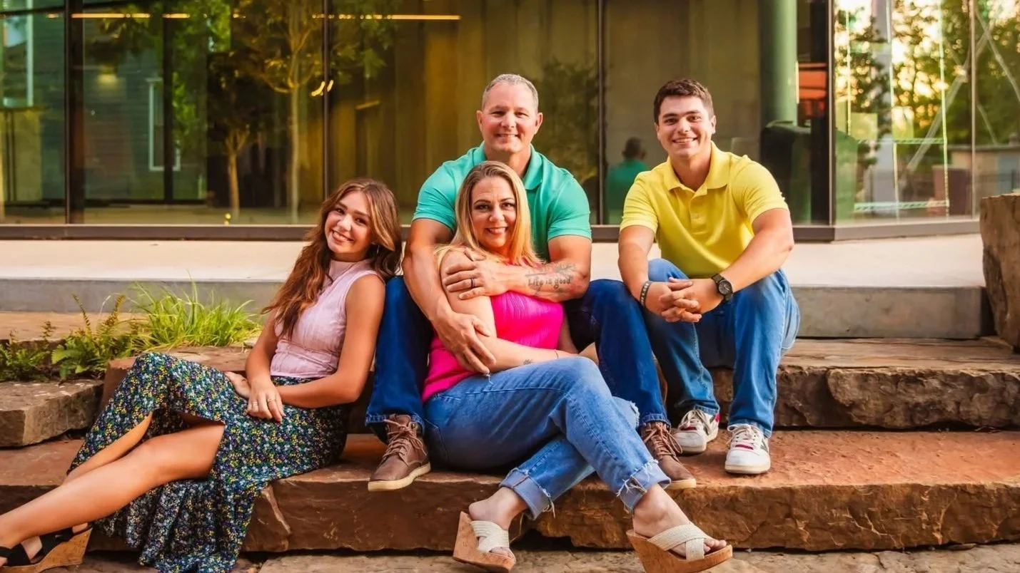 Group of five smiling people sitting outdoors on stone steps, in front of a building with glass windows, during sunset.
