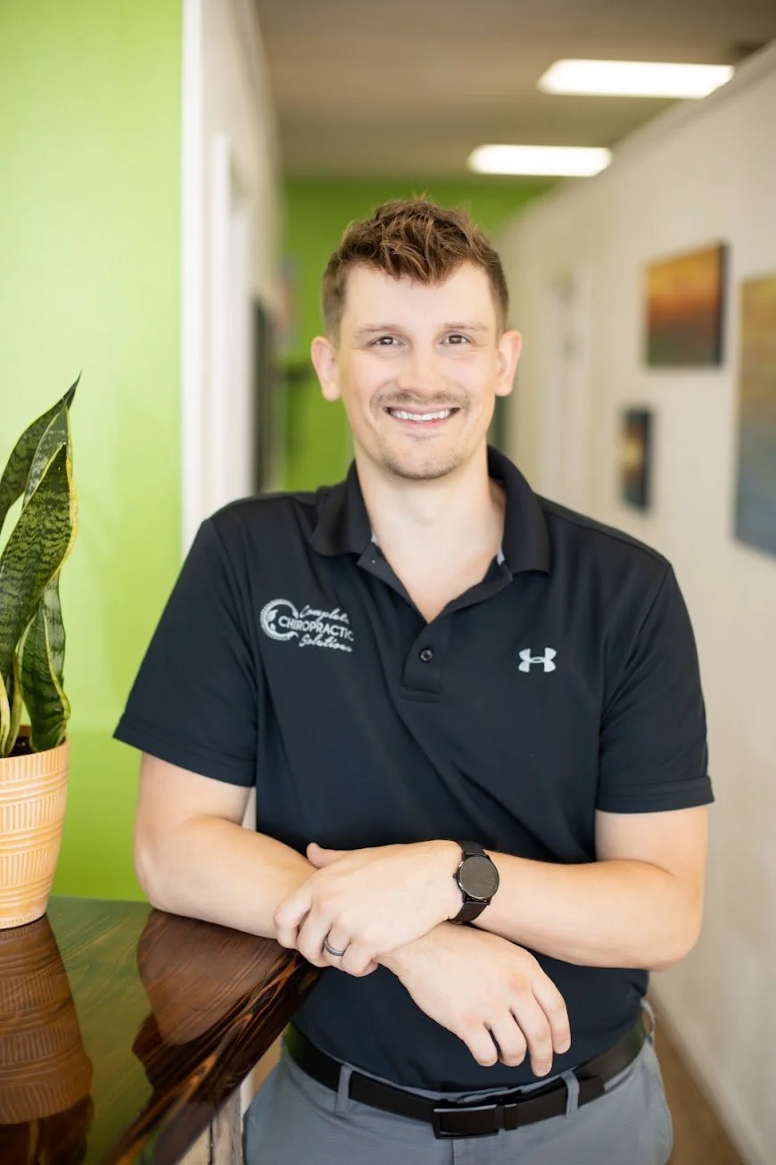 A smiling man with short brown hair wearing a black polo shirt with the logo 'Complete Chiropractic Solutions' and Under Armour logo, standing indoors near a green wall and a potted plant.