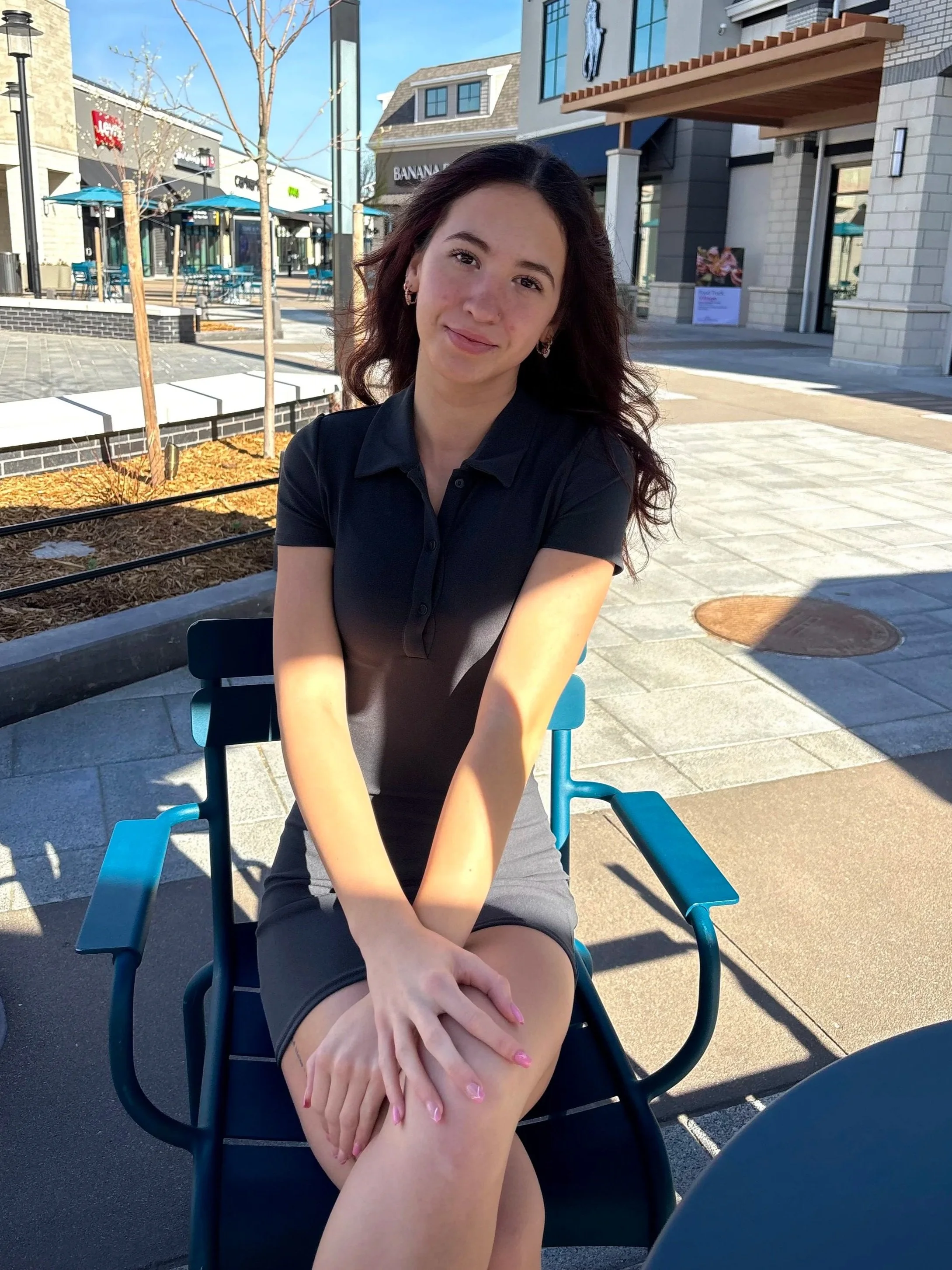 A young woman with dark wavy hair, wearing earrings and a black polo shirt, sitting outdoors on a blue chair at a shopping plaza or outdoor mall during daytime. In the background, there are storefronts, trees, and outdoor seating areas under umbrellas.