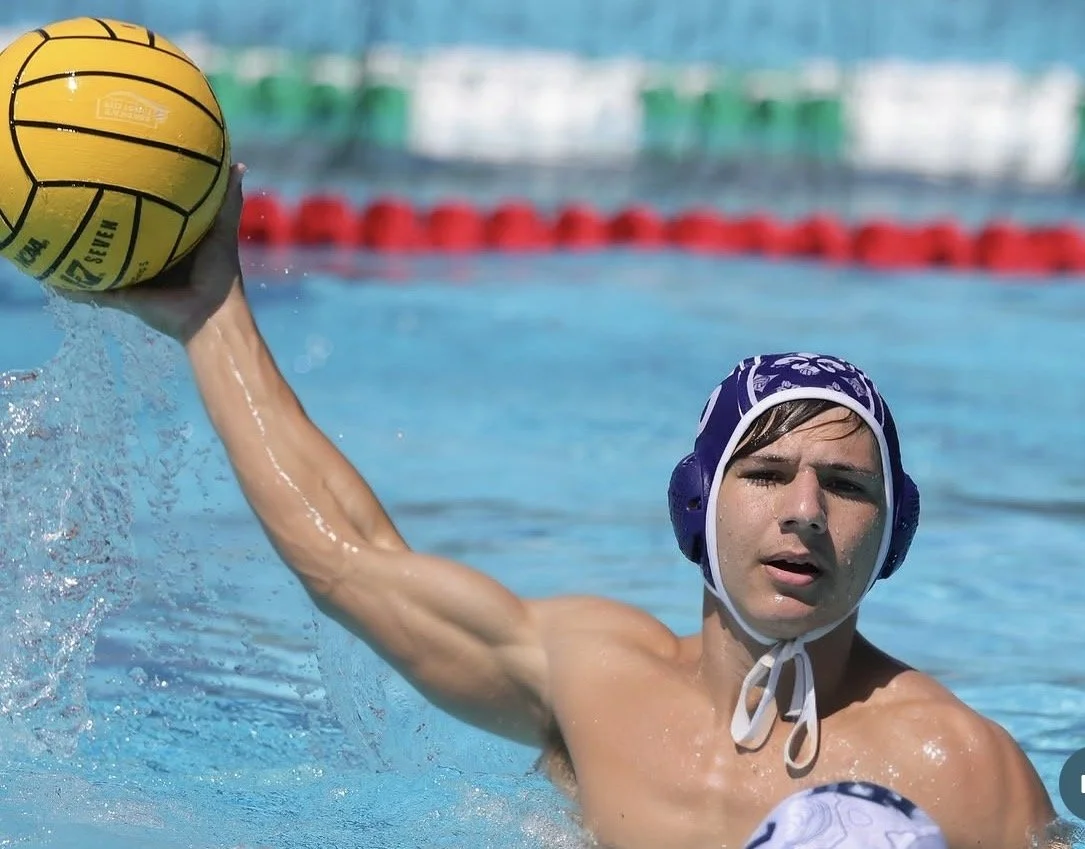 Male water polo player wearing a blue cap with a white chin strap, holding a yellow water polo ball in the water during a game.