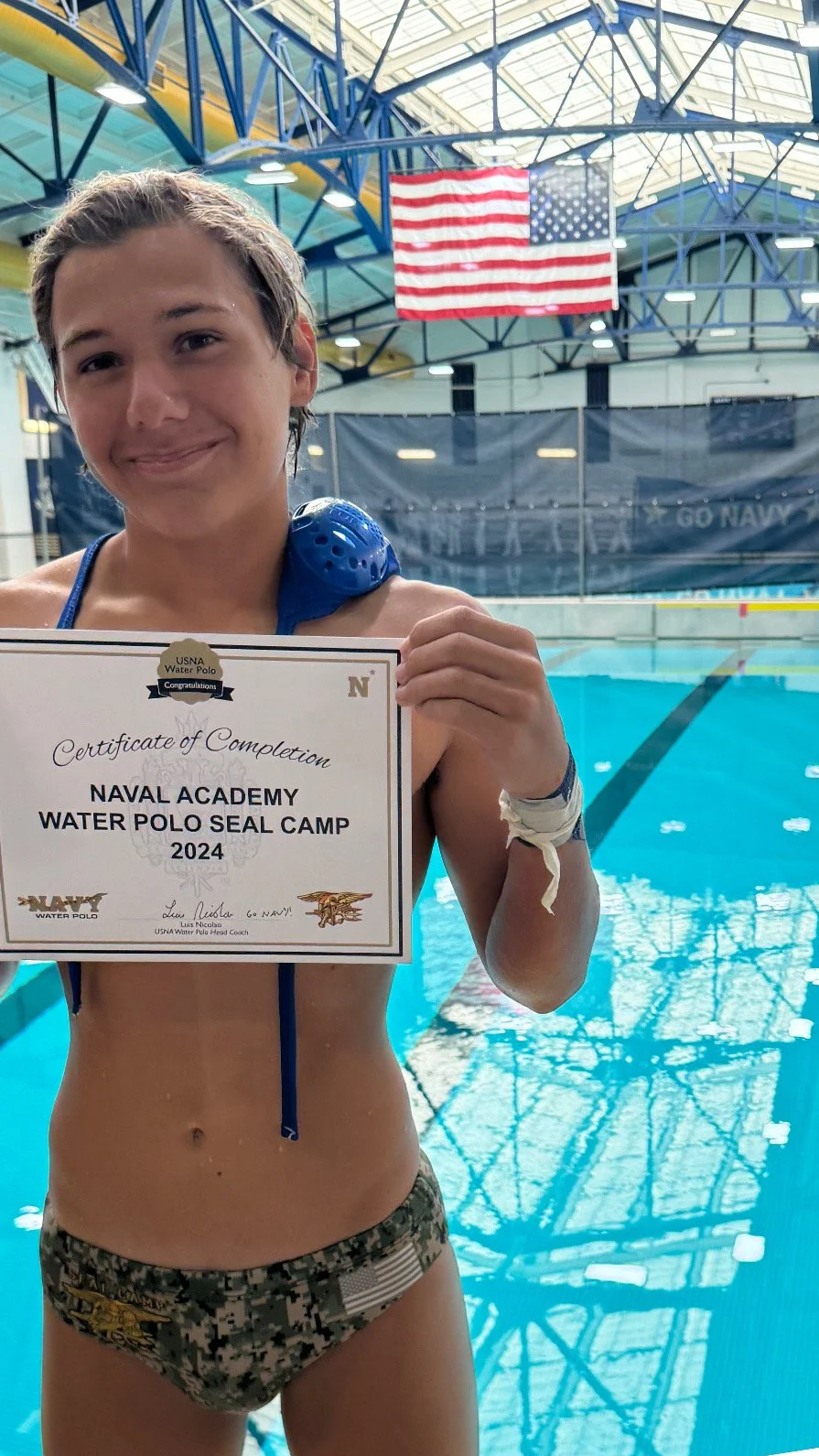 A young woman in water polo attire holding a certificate in an indoor swimming pool facility. She is smiling, with the American flag hanging from the ceiling behind her.