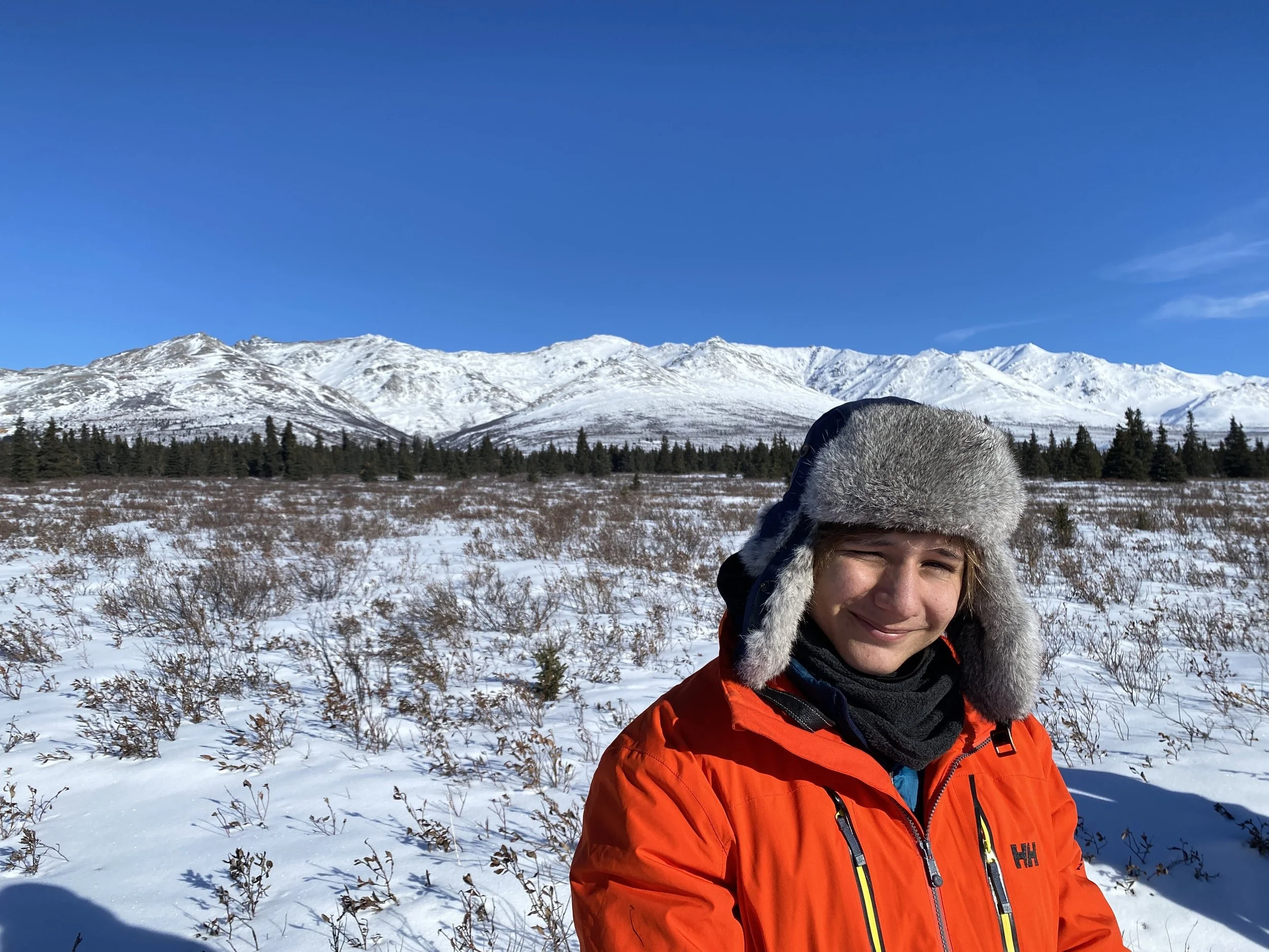Person wearing an orange winter jacket and furry winter hat standing outdoors in a snowy landscape with mountains in the background and clear blue sky.