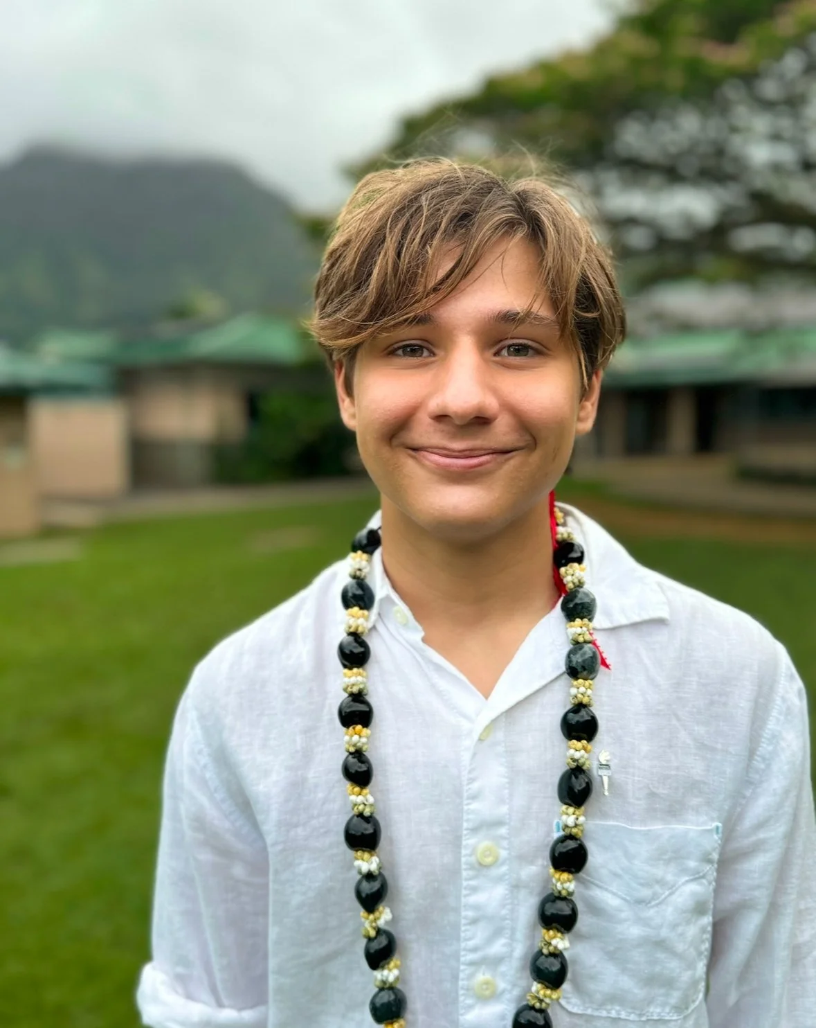 Young man with light brown hair and light skin smiling outdoors, wearing a white button-up shirt and a black and white bead necklace, with a blurred background of grass, trees, and a building.