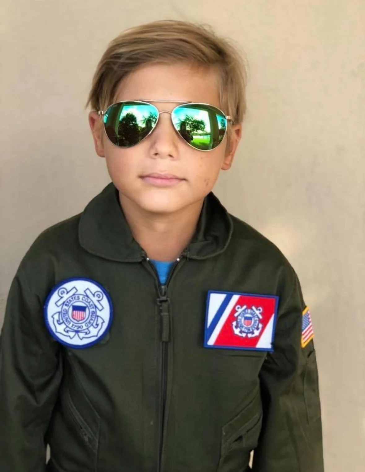 Young boy wearing aviator sunglasses, a green military-style jacket with patches, including a U.S. Coast Guard PATCH, a fire department patch, and an American flag PATCH, standing against a plain beige wall.
