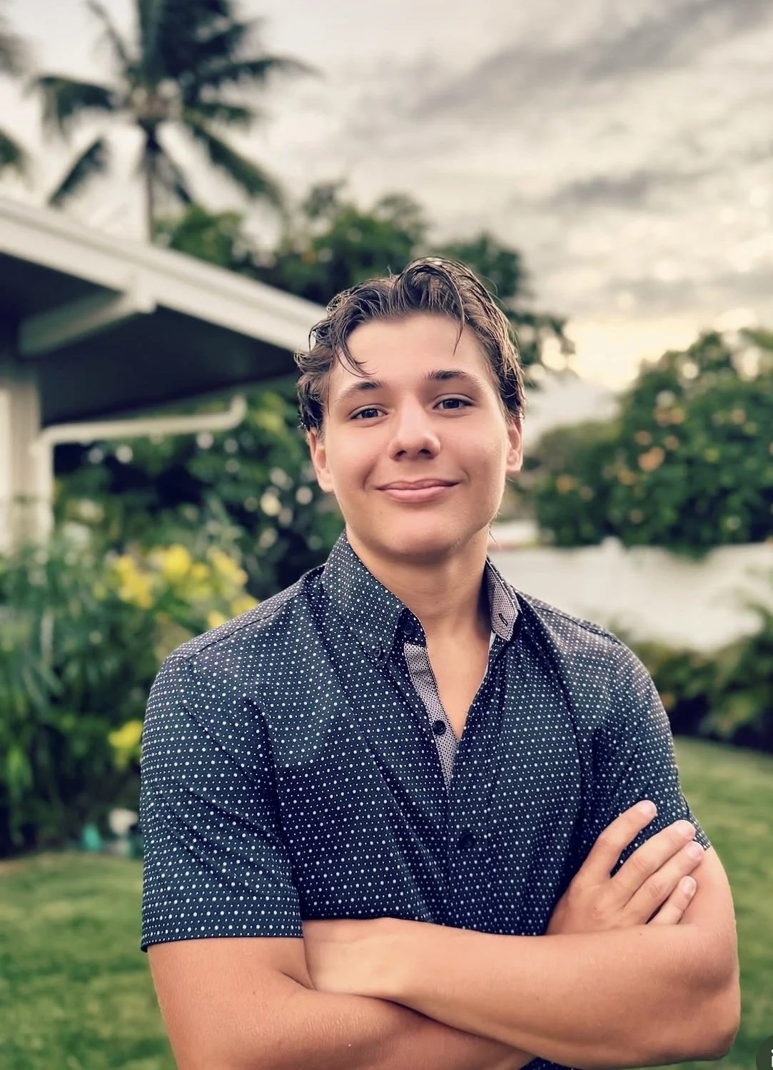 A young man with wavy brown hair smiling in a garden with trees, grass, and a house in the background.
