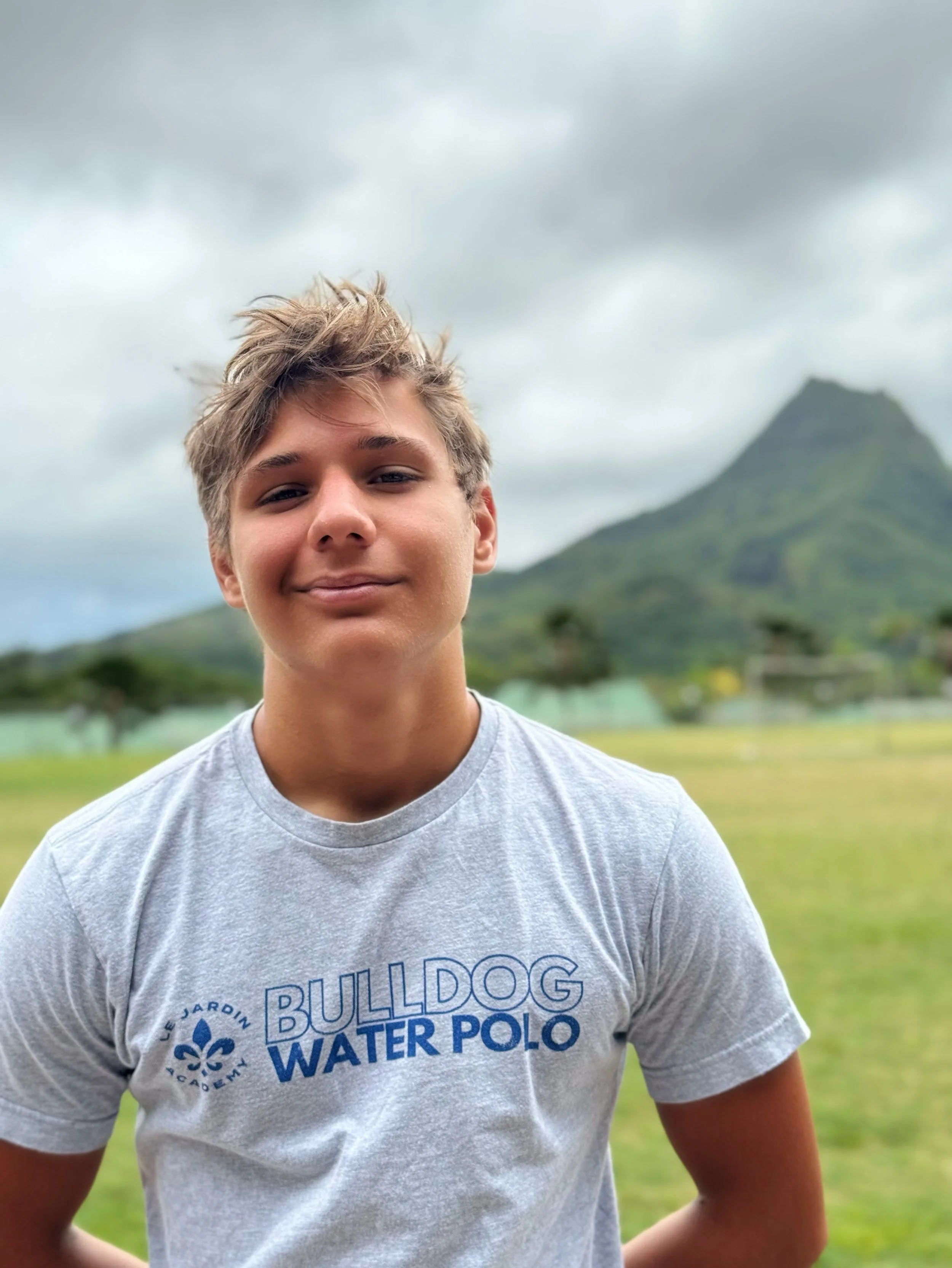 Close-up of a smiling young man with tousled hair wearing a gray T-shirt with blue text and logo, standing outdoors with a mountain and cloudy sky in the background.