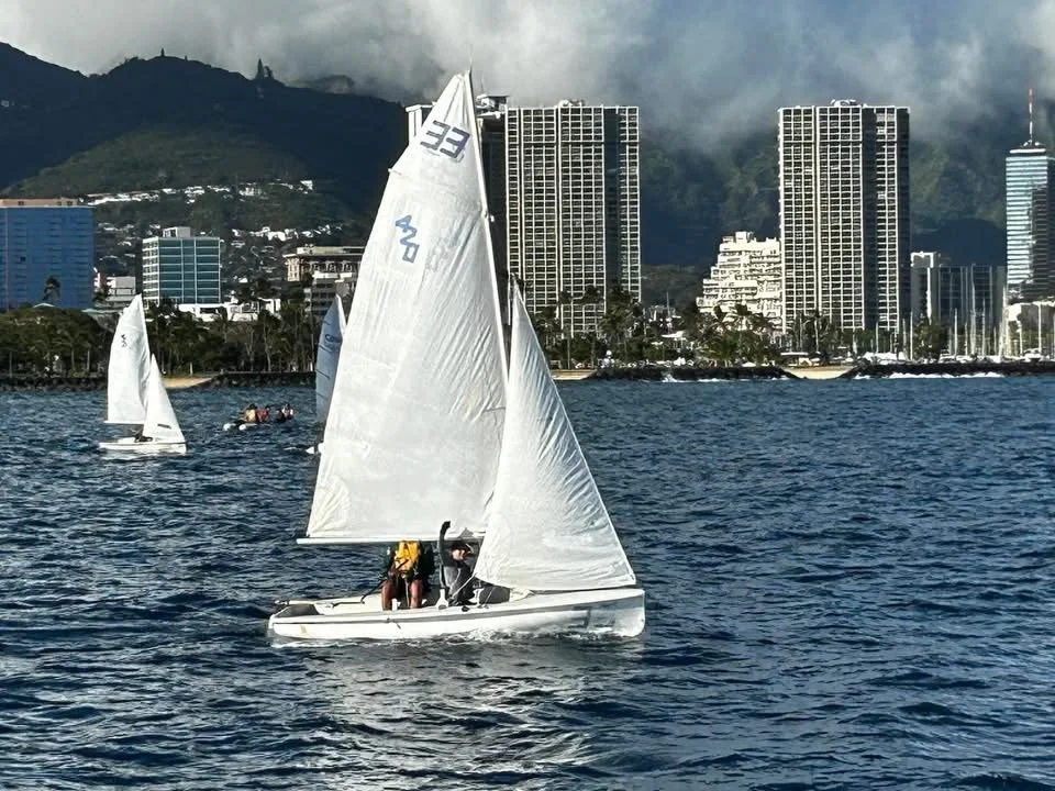 Sailing boat with two people sailing on the water in front of a city skyline with high-rise buildings and mountains in the background.