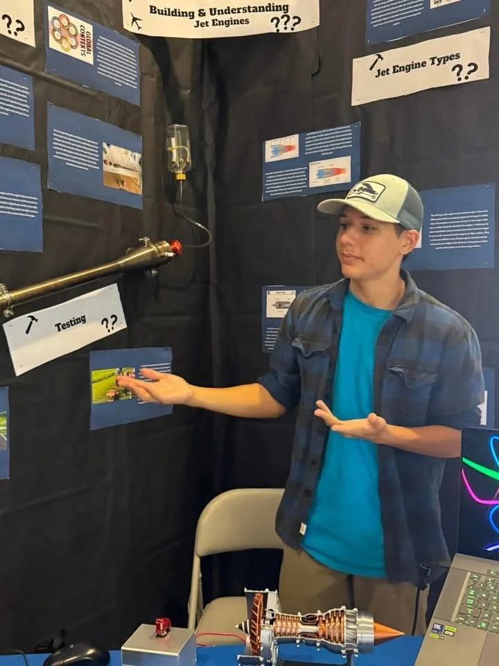 A young man in a blue t-shirt, plaid shirt, and baseball cap standing next to a display about jet engines, with informational posters and a test setup with engine testing equipment and a laptop. He is explaining or presenting the display.