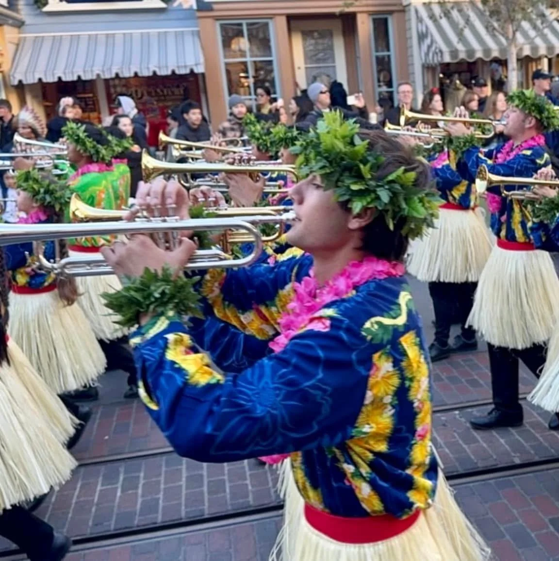 Young people in traditional Hawaiian attire playing musical instruments during a parade, with onlookers in the background.