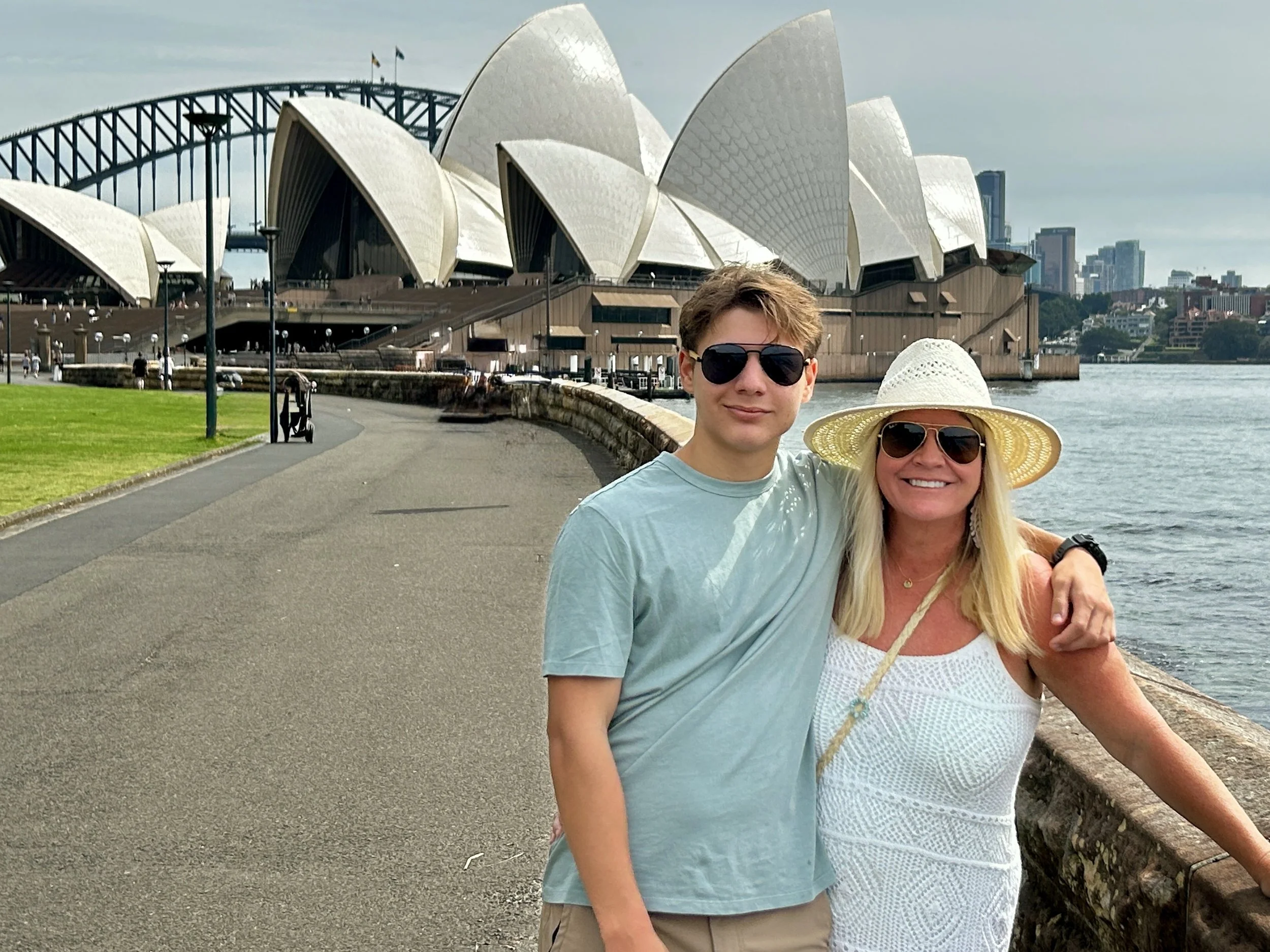Two people are standing together near the Sydney Opera House with the Sydney Harbour Bridge in the background. The man has short brown hair, is wearing dark sunglasses, a light blue T-shirt, and beige shorts. The woman has long blonde hair, is wearing a white sun hat, dark sunglasses, and a white sleeveless top. They are smiling and posing for the photo by the waterfront.