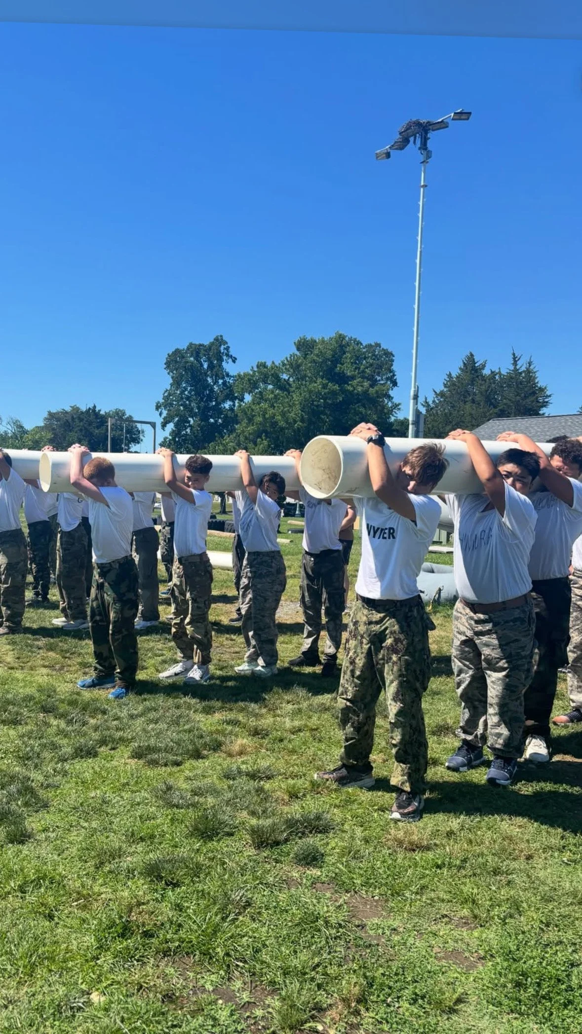 Group of young people in army camouflage pants and white T-shirts, carrying a long white tube outdoors on a grassy field on a sunny day.